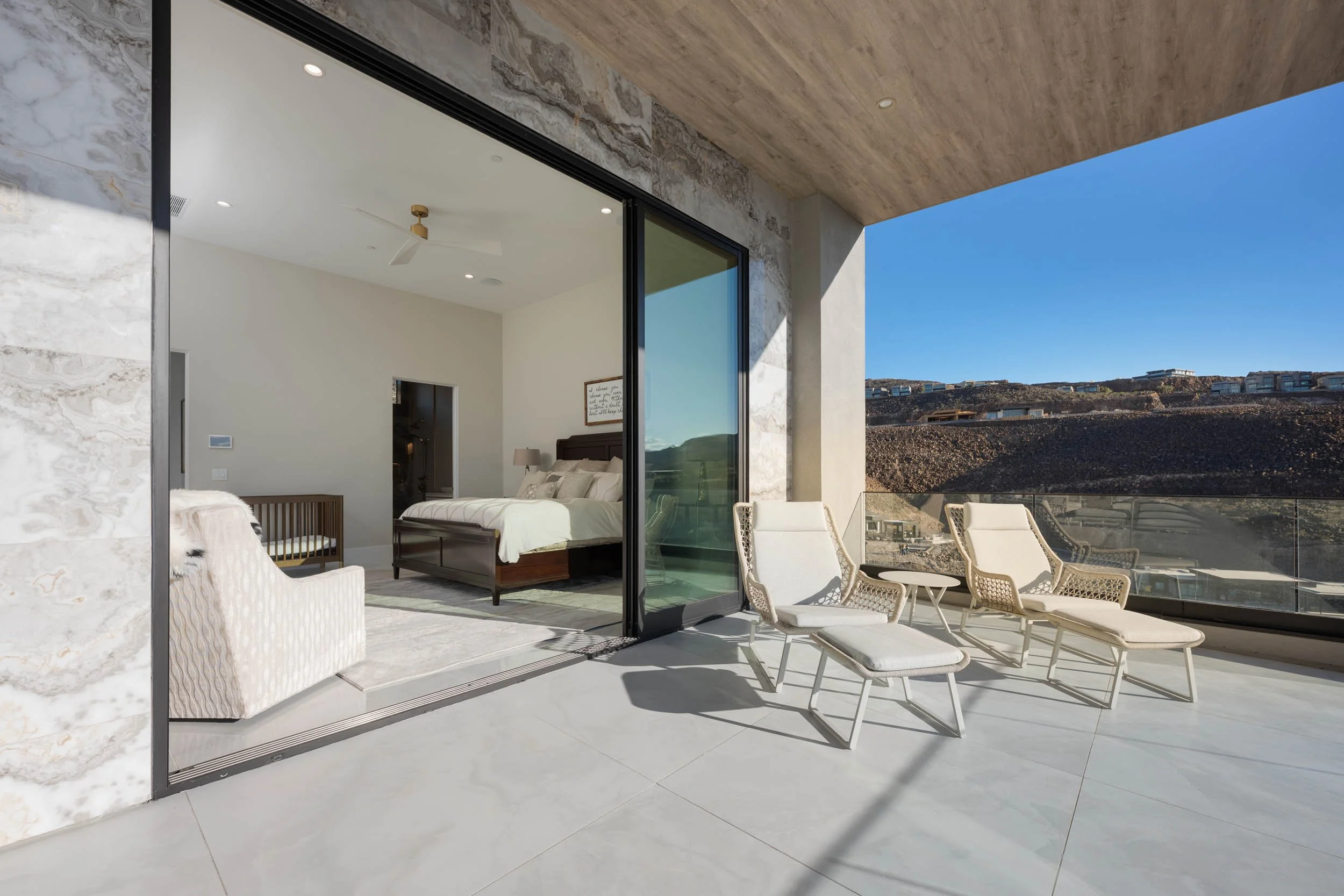 Modern bedroom with large sliding glass door opening to a balcony with beige lounge chairs, scenic hillside view, and a blue sky.