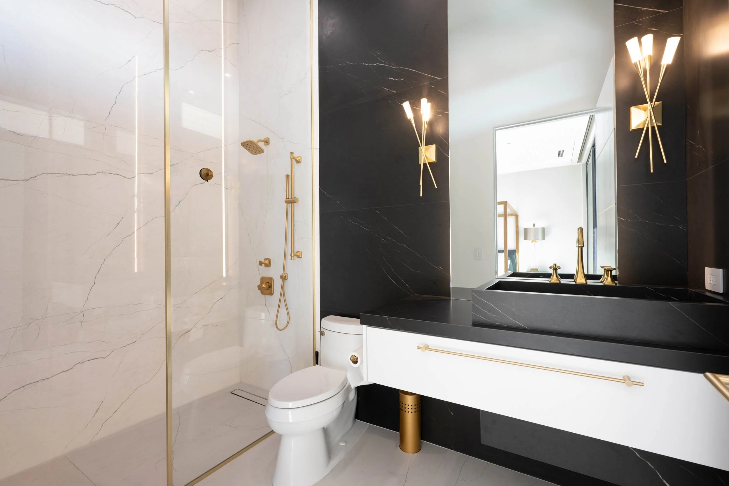 Modern bathroom with black and white marble walls, gold fixtures, a walk-in shower, white toilet, black countertop with a white cabinet, large mirror, and stylish wall-mounted light fixtures.