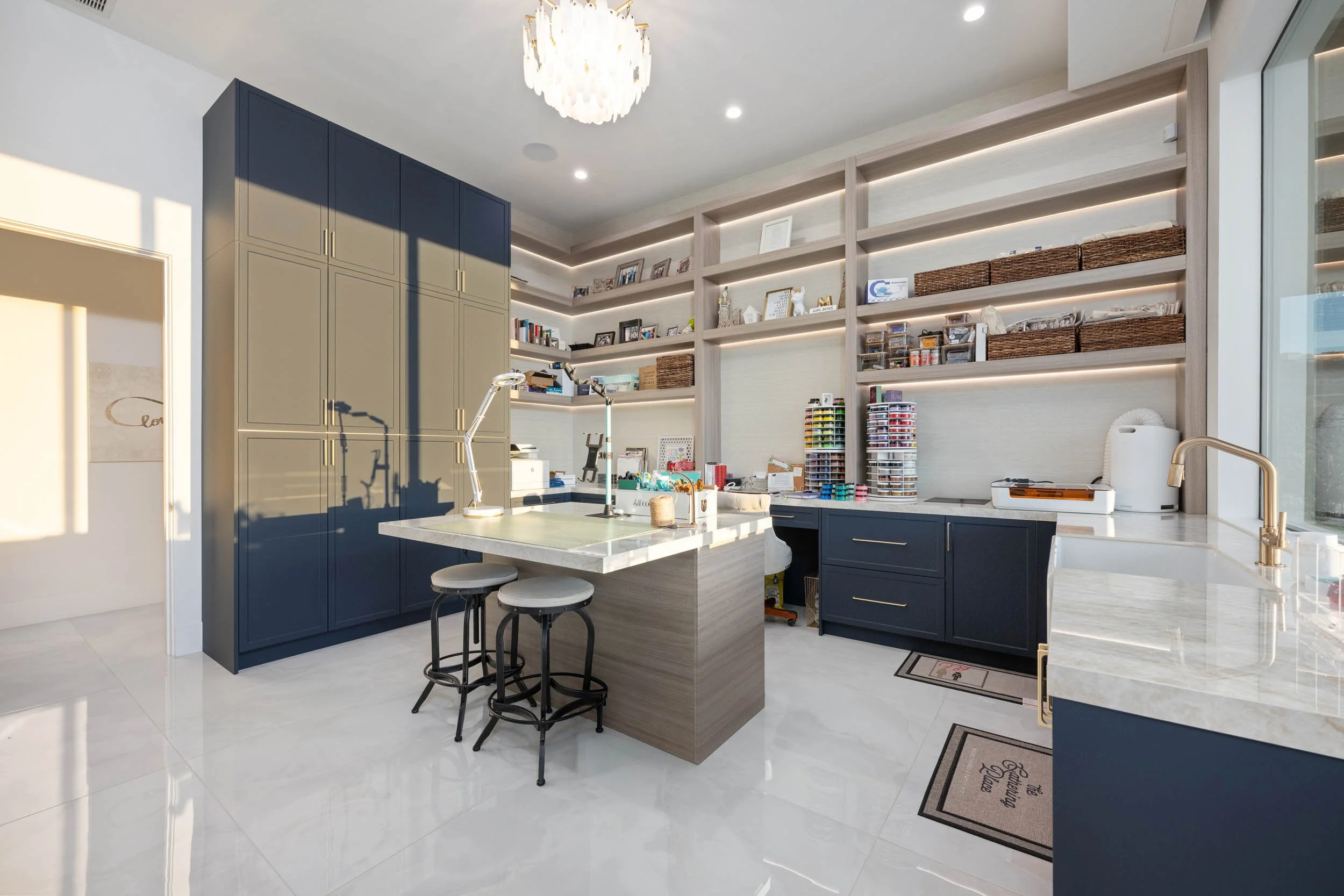 A modern kitchen with navy blue cabinets, a white marble island with seating, open shelving with organization supplies, a large window, and a chandelier lighting fixture.