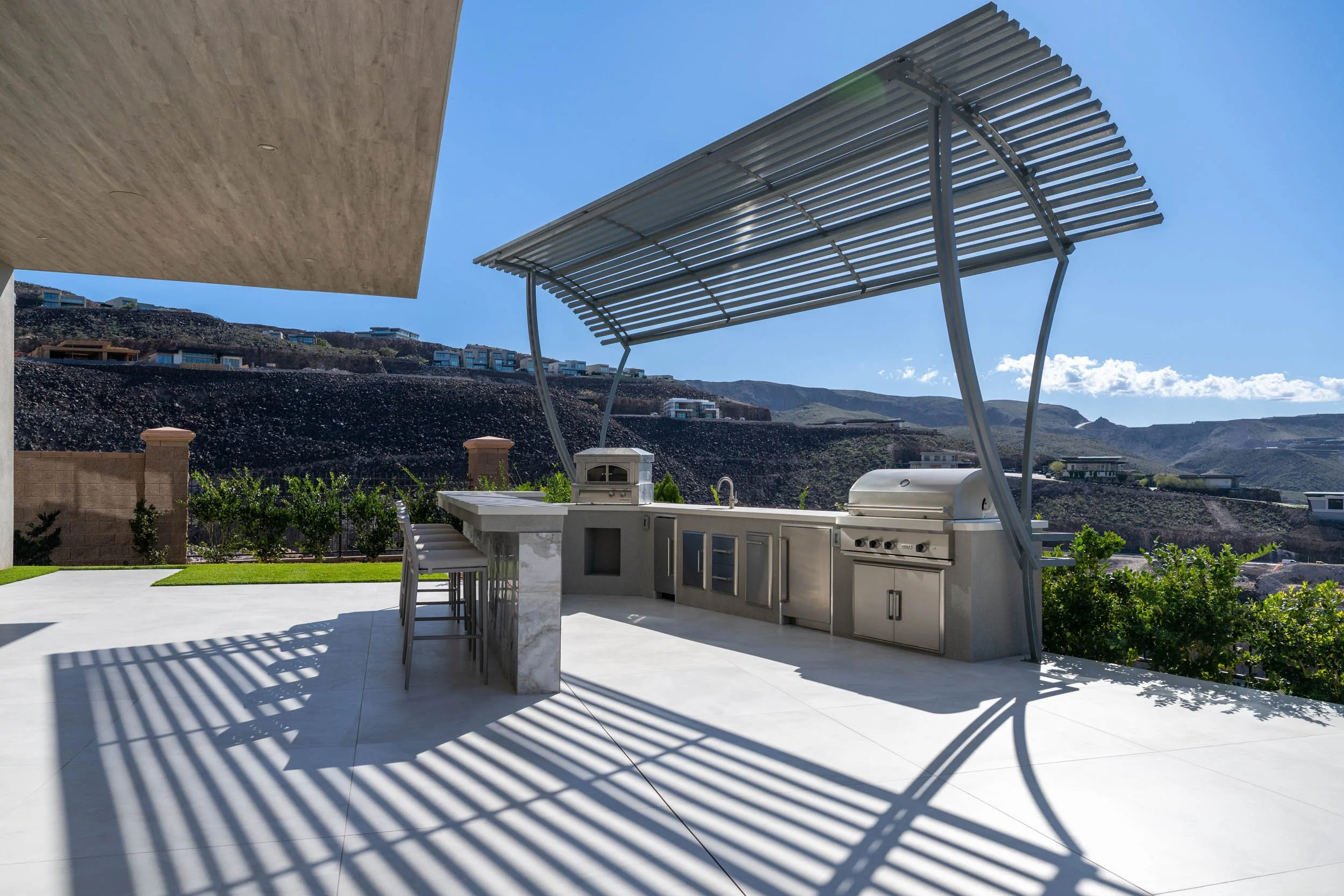 Outdoor kitchen area with a stainless steel barbecue grill, a countertop, and a shaded pergola, overlooking a hilly landscape under a clear blue sky.
