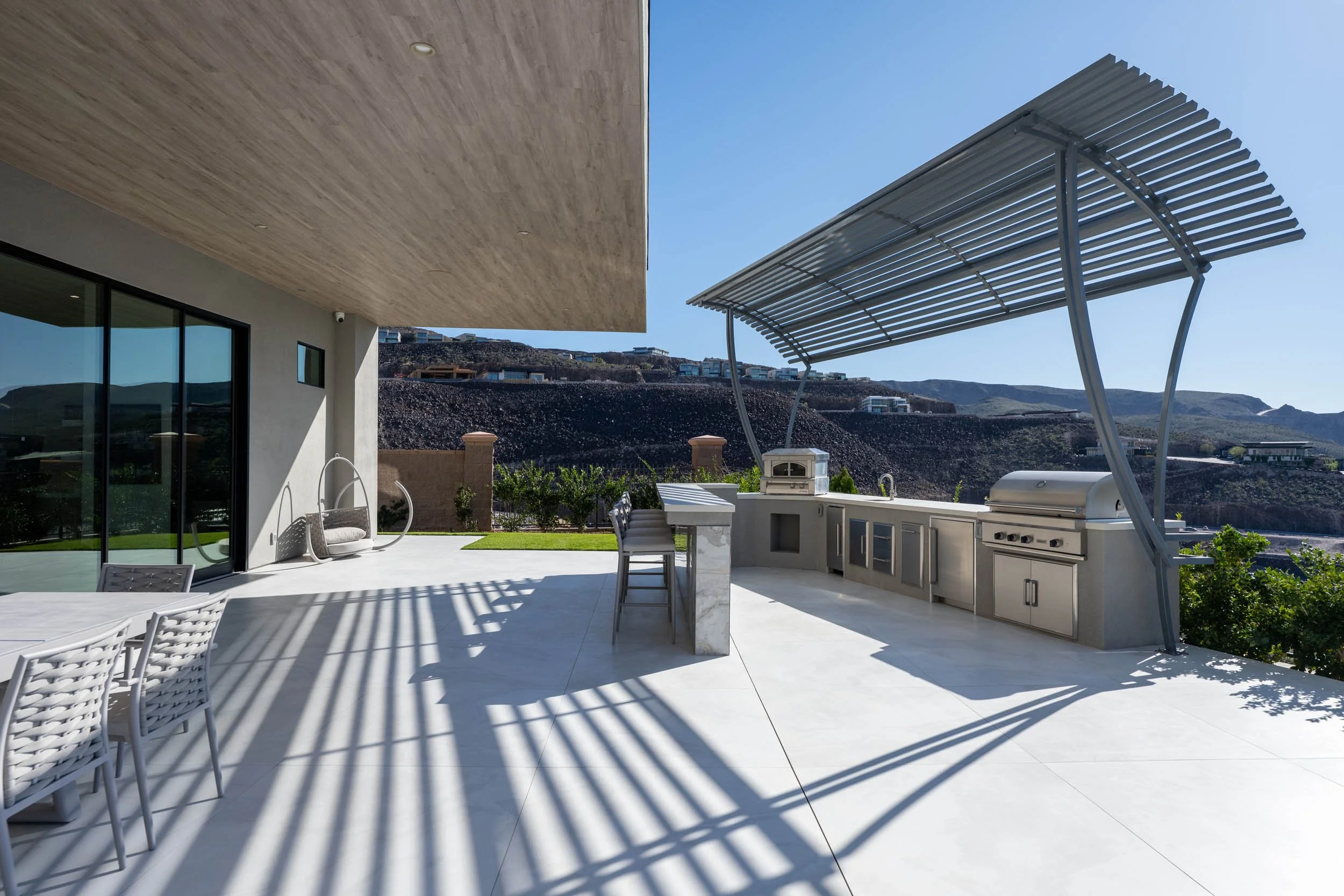 Modern outdoor patio with an outdoor kitchen and grill, shaded by a curved metal awning; shadow patterns cast on the white tiled floor, overlooking mountains and houses in the distance.