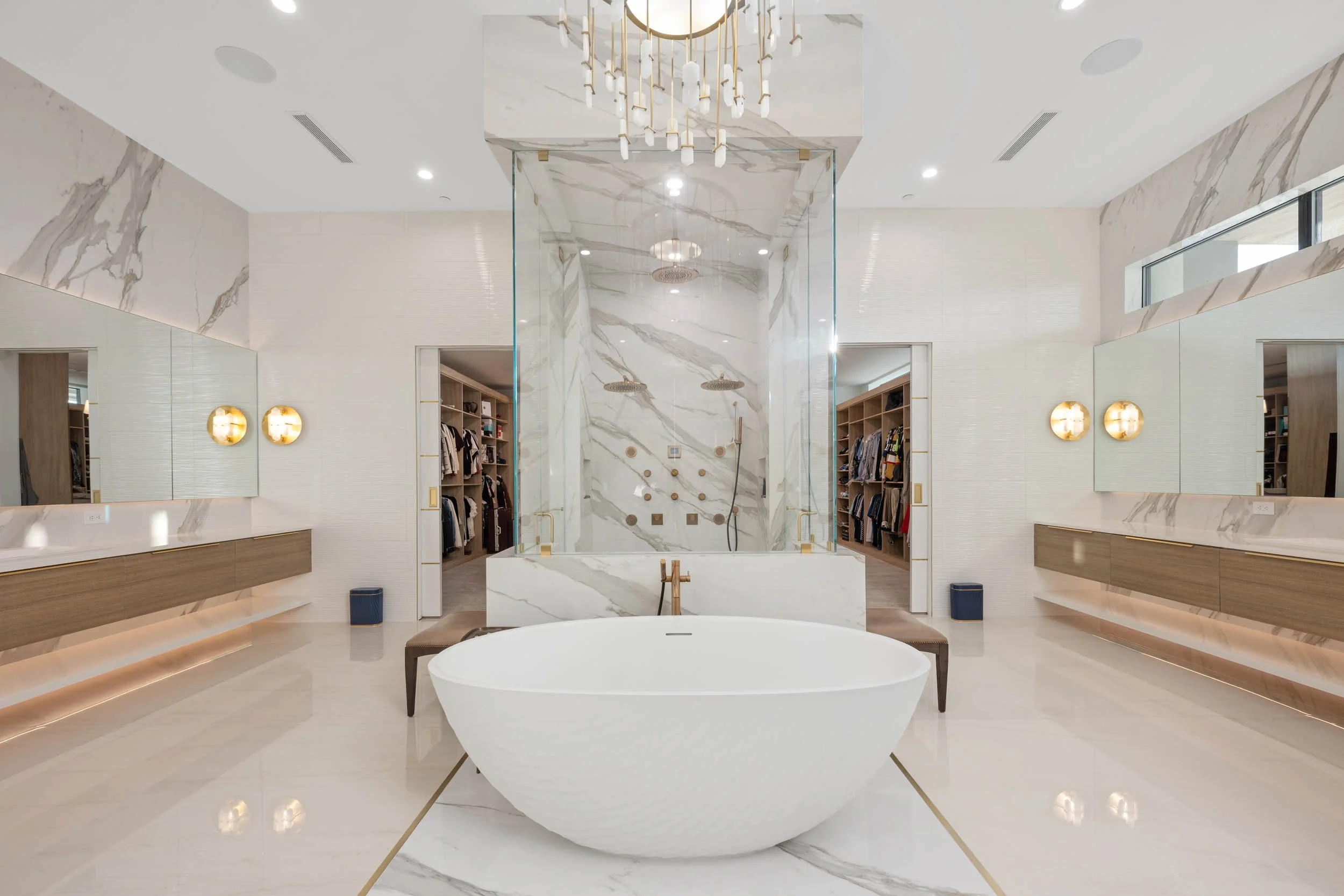 A luxurious modern bathroom with a freestanding bathtub, marble walls, and a glass-enclosed shower in the background. There are wide mirrors and floating vanities on both sides, with warm lighting and a chandelier hanging from the ceiling.