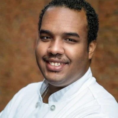 Portrait of a man with short curly black hair, smiling, wearing a white chef's coat, against a brown brick wall background.