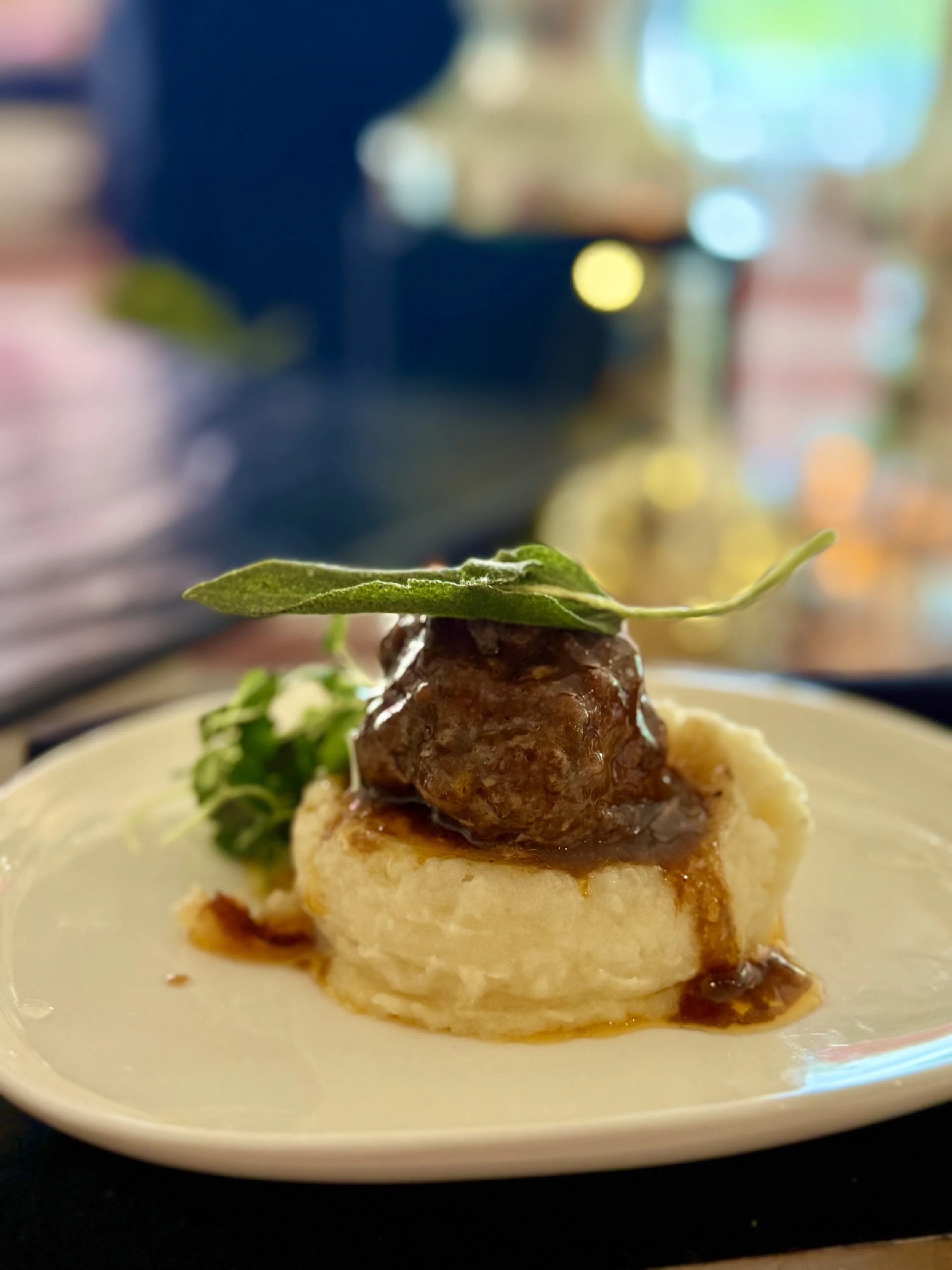 A plated dish with mashed potatoes topped with a meatball covered in sauce, garnished with a fresh green herb, on a white plate with a blurred colorful background.