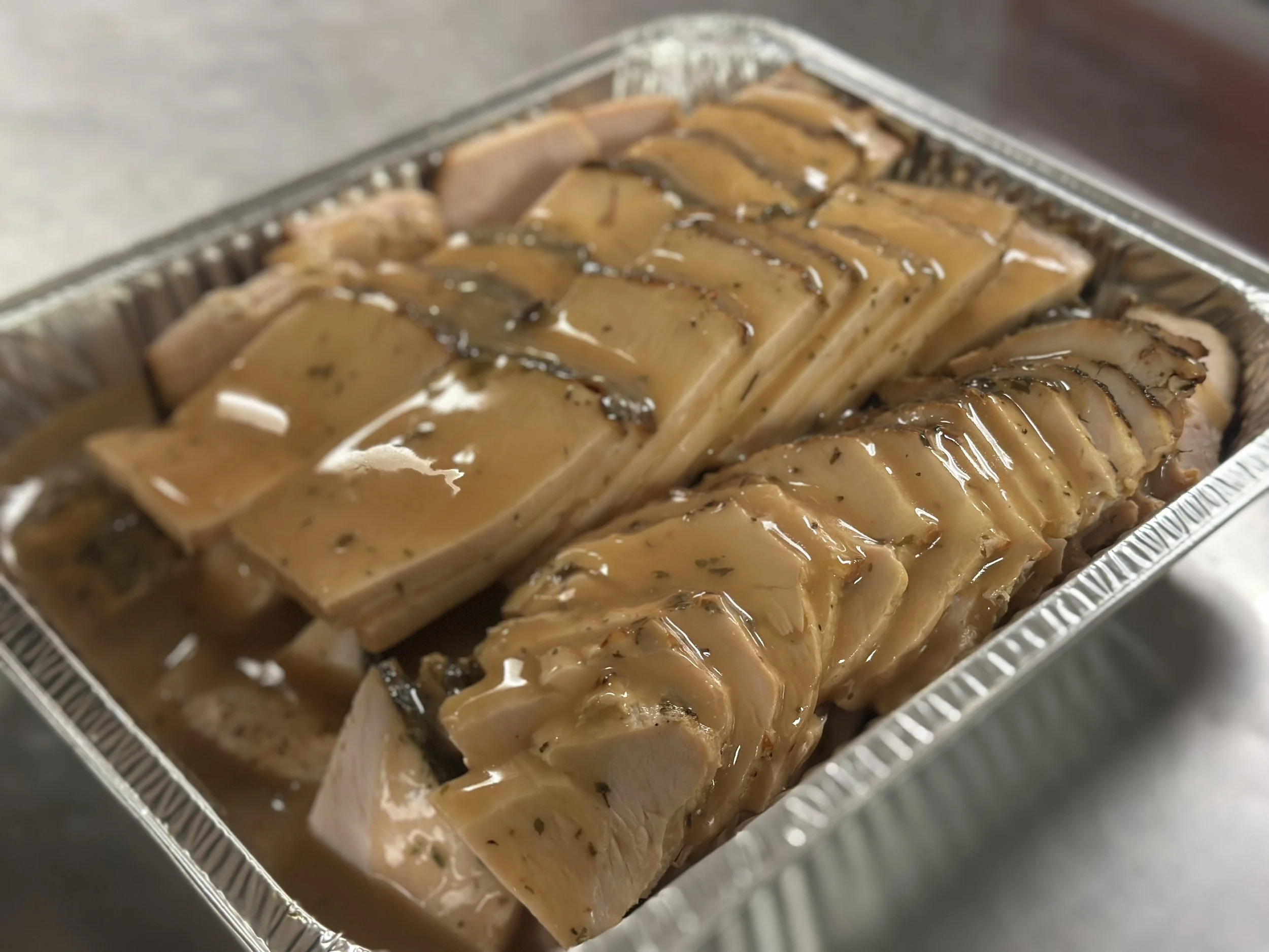 Sliced cooked meat, possibly ham or pork, topped with brown gravy in a foil tray.