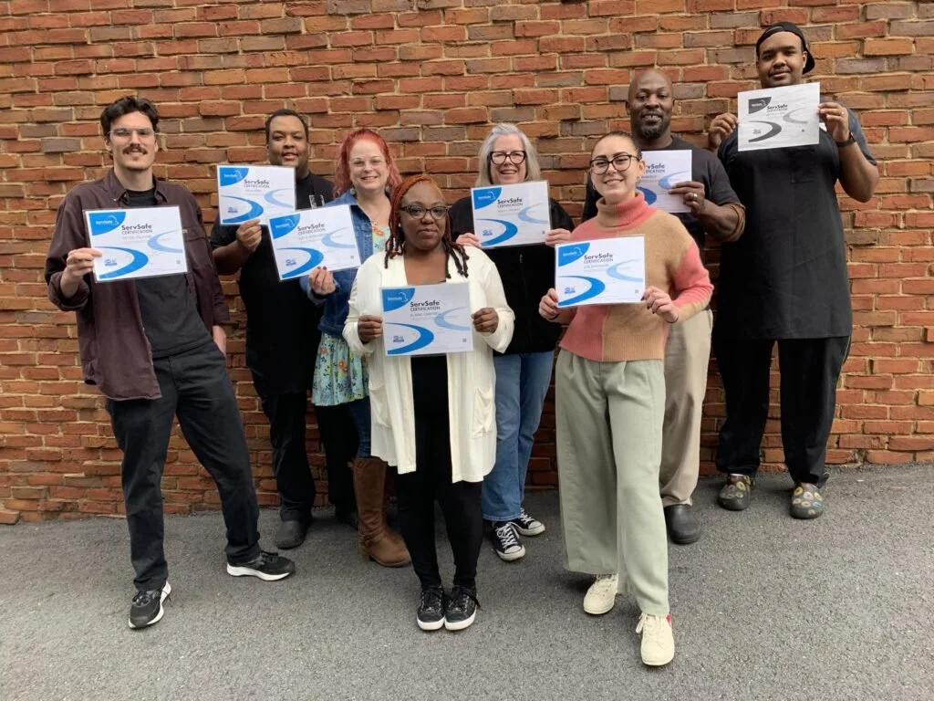 Group of eight diverse people standing against a brick wall, holding certificates that read 'ServSafe'.