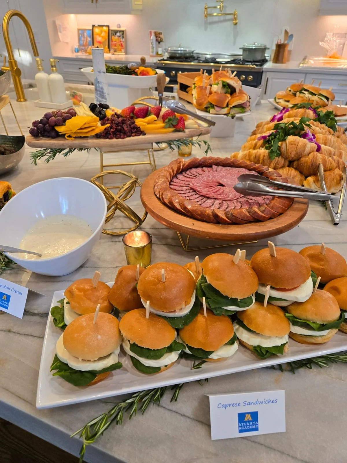 A buffet table with assorted foods including mini caprese sandwiches, sliced cured meats, fresh fruit, croissants, and sandwiches, with utensils and small decorative items.