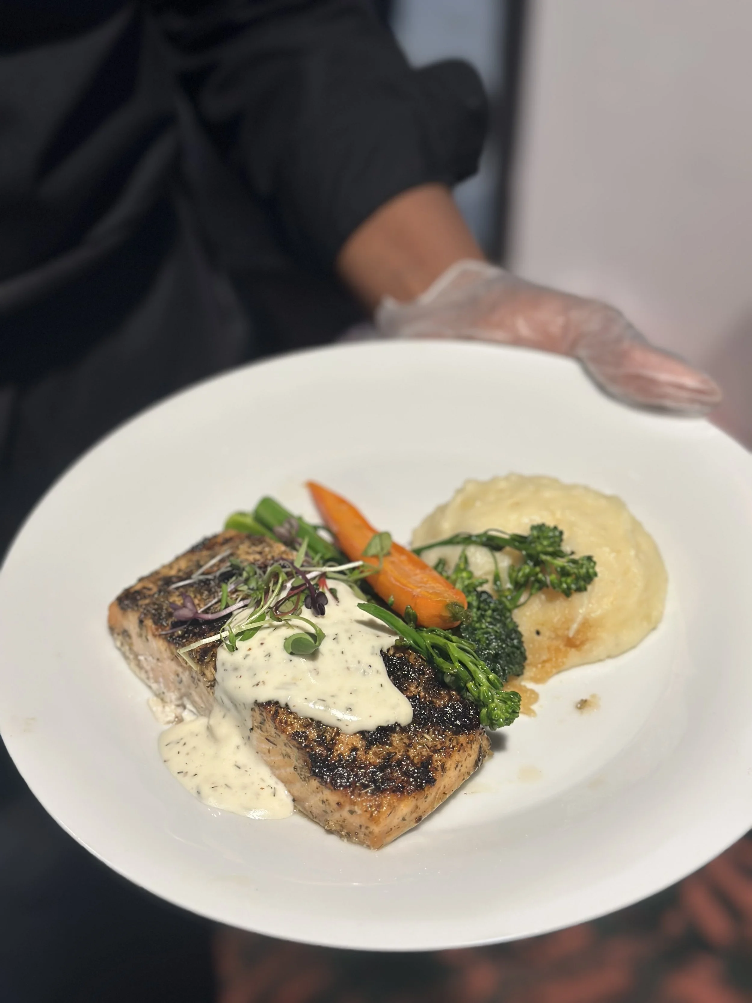 Plate with grilled salmon topped with creamy sauce, garnished with microgreens, served with mashed potatoes and mixed vegetables including carrots and broccoli.