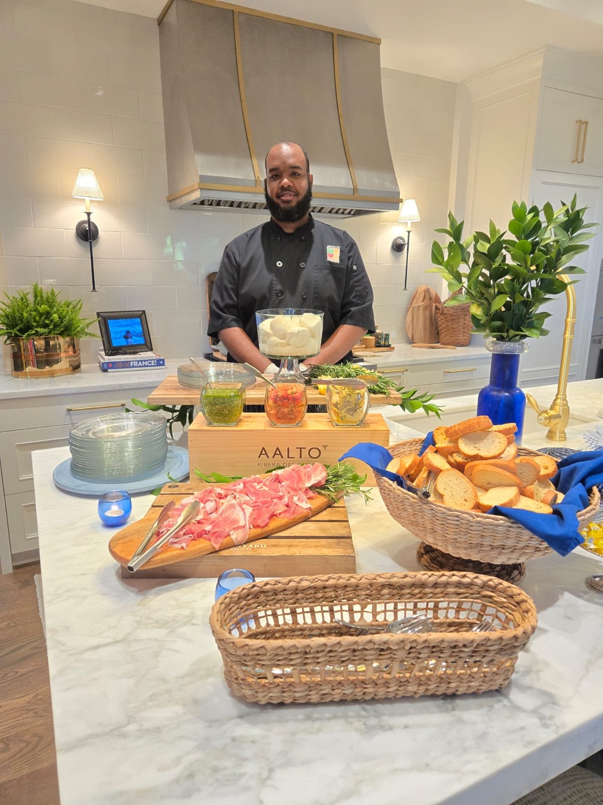 A man in a black chef uniform standing behind a kitchen counter with various food ingredients and dishes, including sliced bread in a basket, sliced cured meat on a wooden board, jars of pesto and tomato relish, and a large bowl of mozzarella balls. There are decorative plants, framed photos, and kitchen appliances in the background.