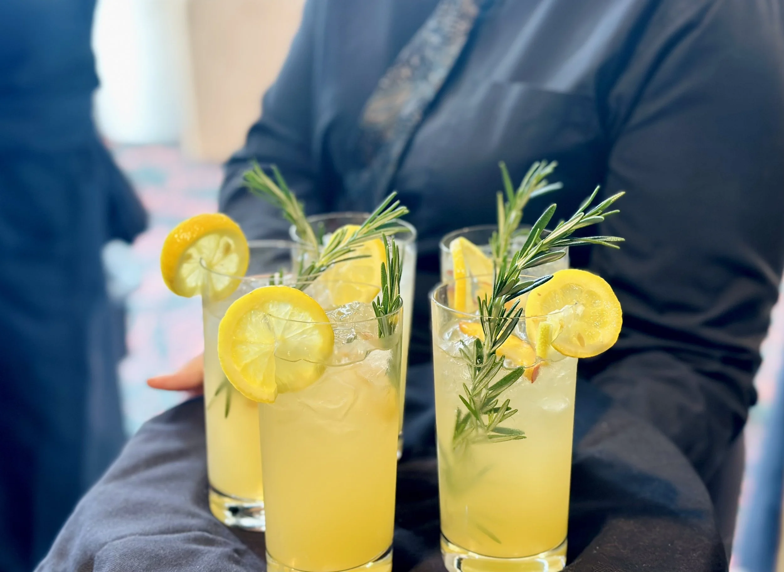 Several tall glasses of lemonade garnished with slices of lemon and sprigs of rosemary, held by a person in a dark jacket.