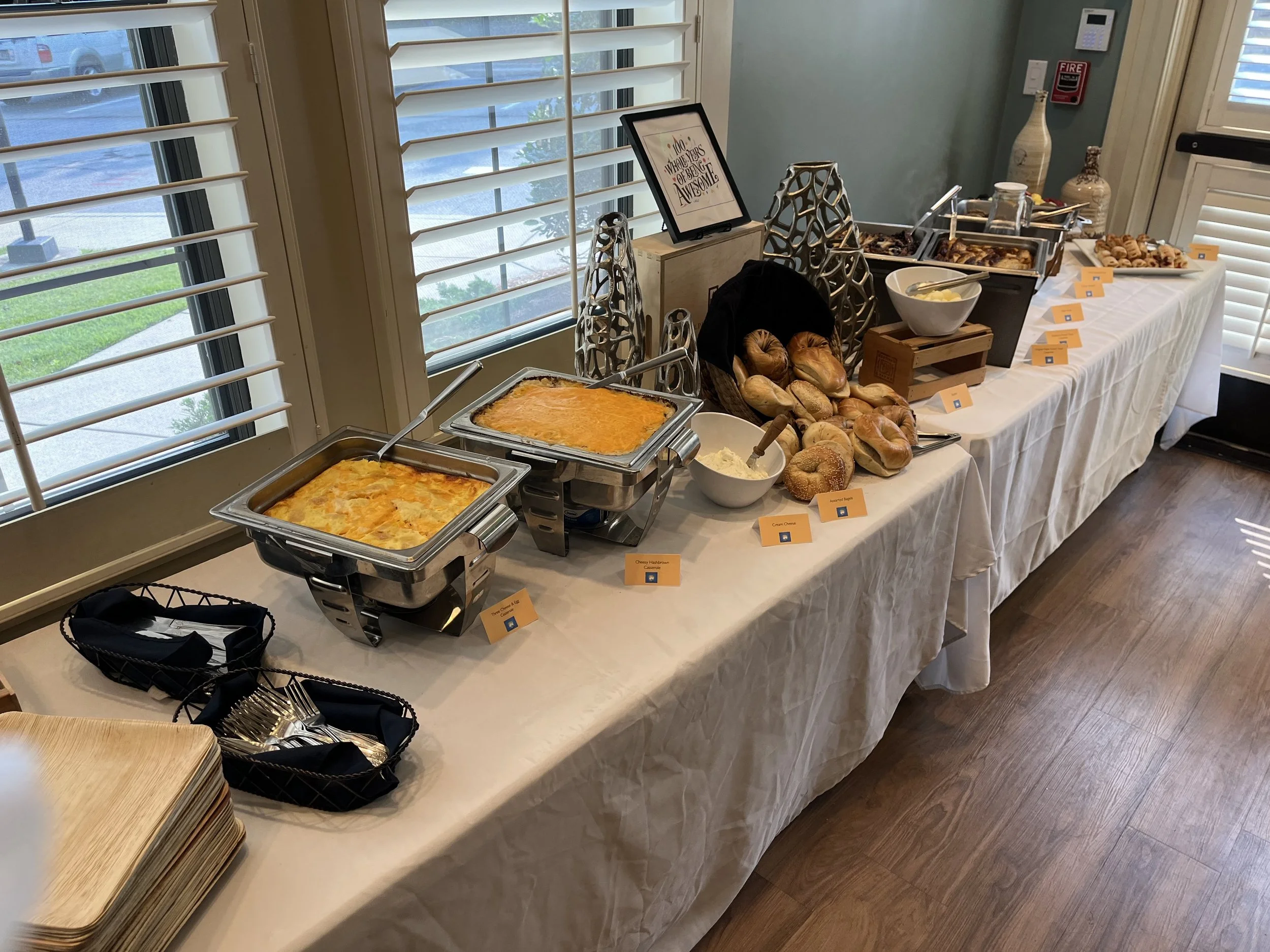 A buffet table with breakfast items including baked goods, scrambled eggs, and toppings, set up near windows with white shutters and a green wall.