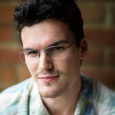 A young man with dark hair and glasses, wearing a light-colored shirt, smiling slightly at the camera with a brick wall background.