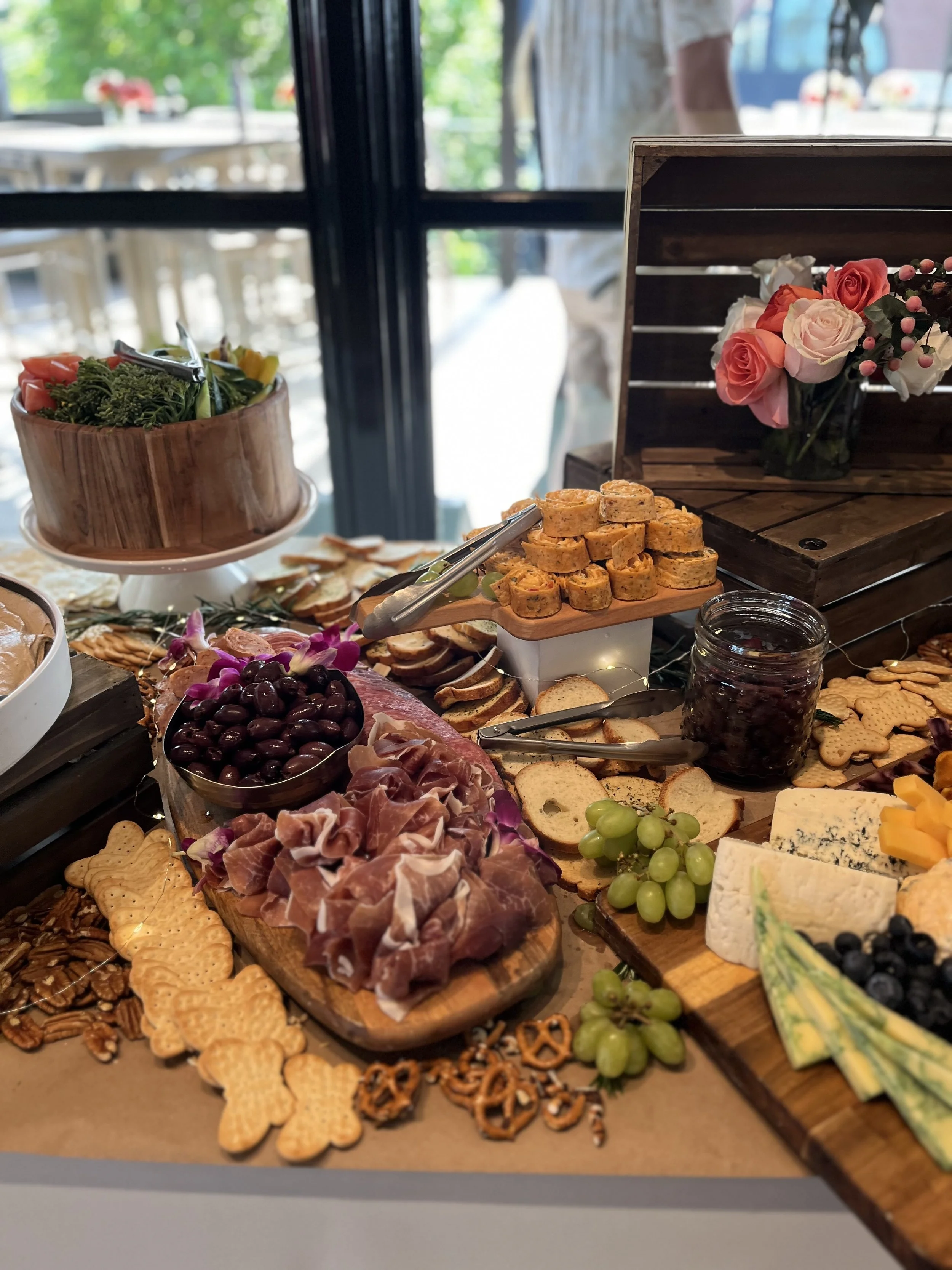 A spread of appetizers on a buffet table, including cheese, grapes, crackers, sliced meats, and a salad. There is a vase with pink and white roses and a wooden box with utensils. The background shows a window with a blurred outdoor scene.