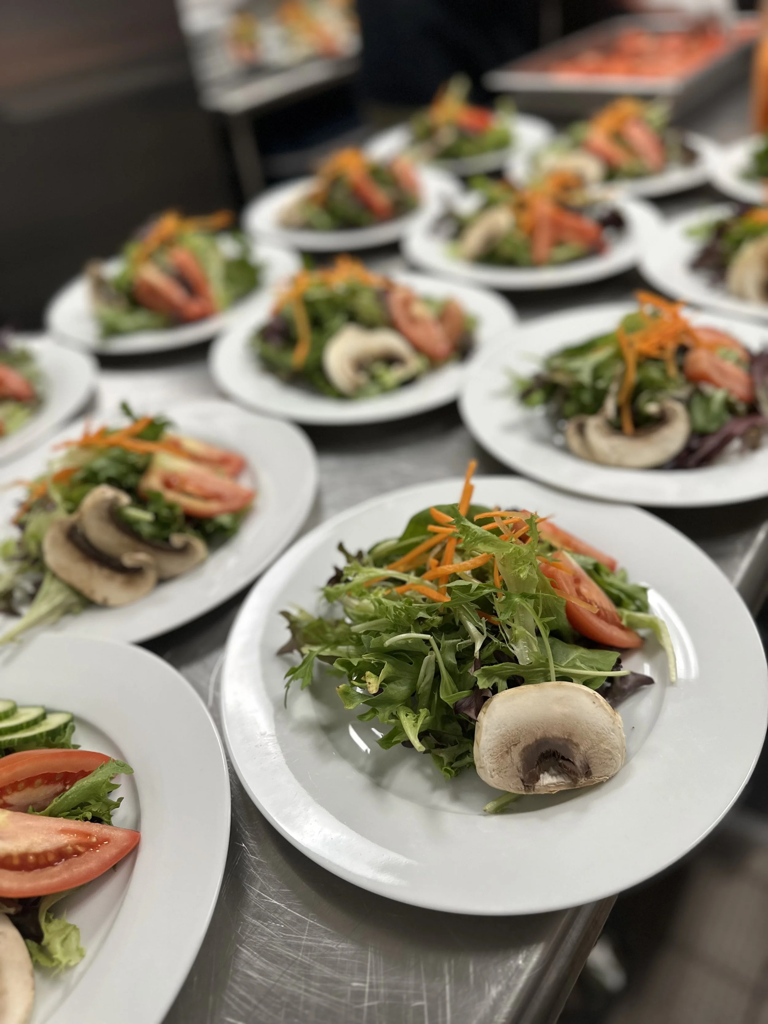 Plates of fresh salad with mixed greens, tomatoes, carrots, cucumber, and mushrooms, arranged on a stainless steel countertop.
