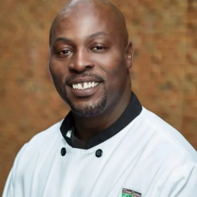 A smiling man with dark skin, a shaved head, and a short beard, wearing a white chef's coat, standing in front of a brick wall.
