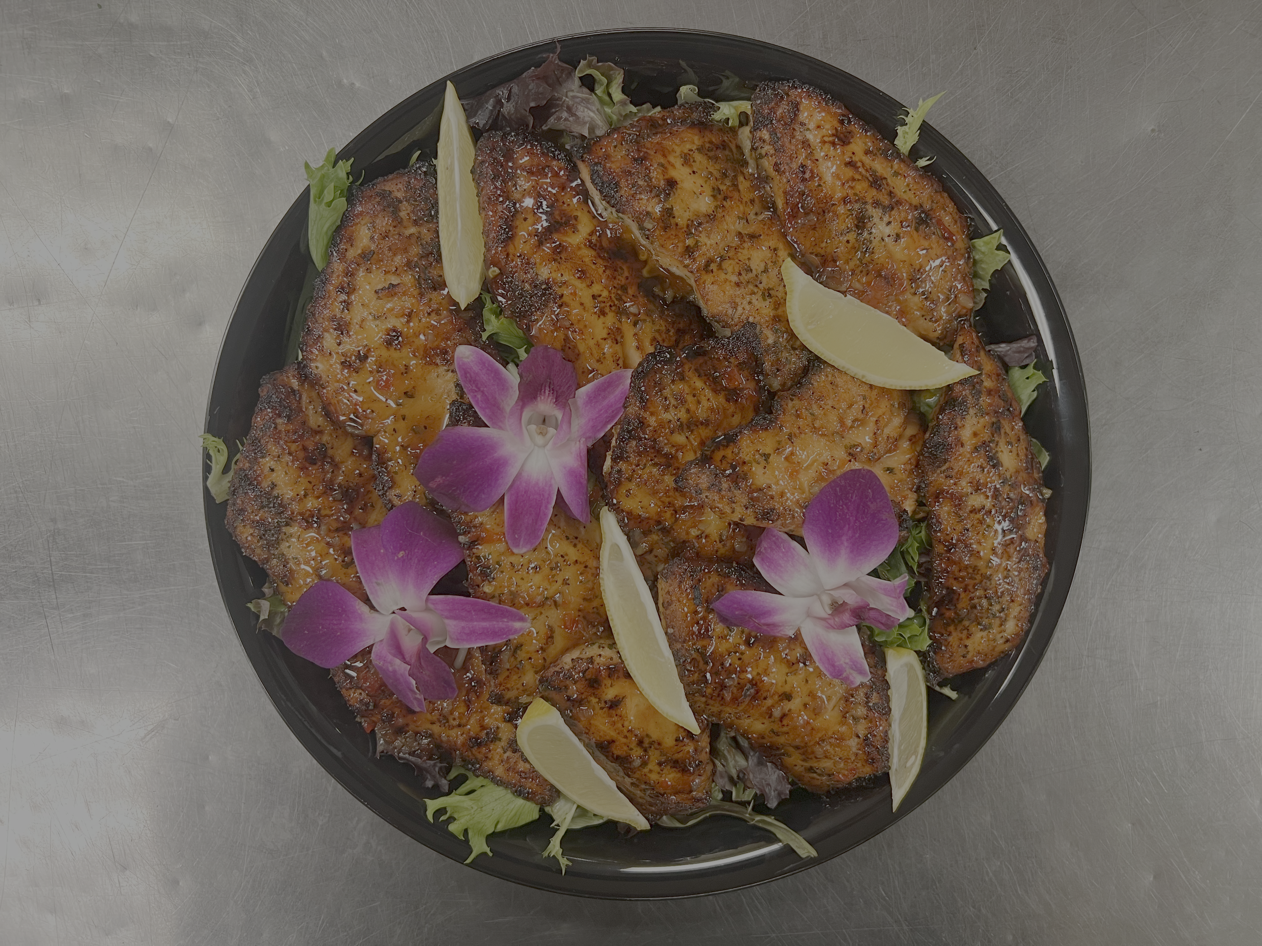 Grilled chicken topped with a glaze, garnished with lemon wedges, edible flowers, and fresh greens on a black platter.