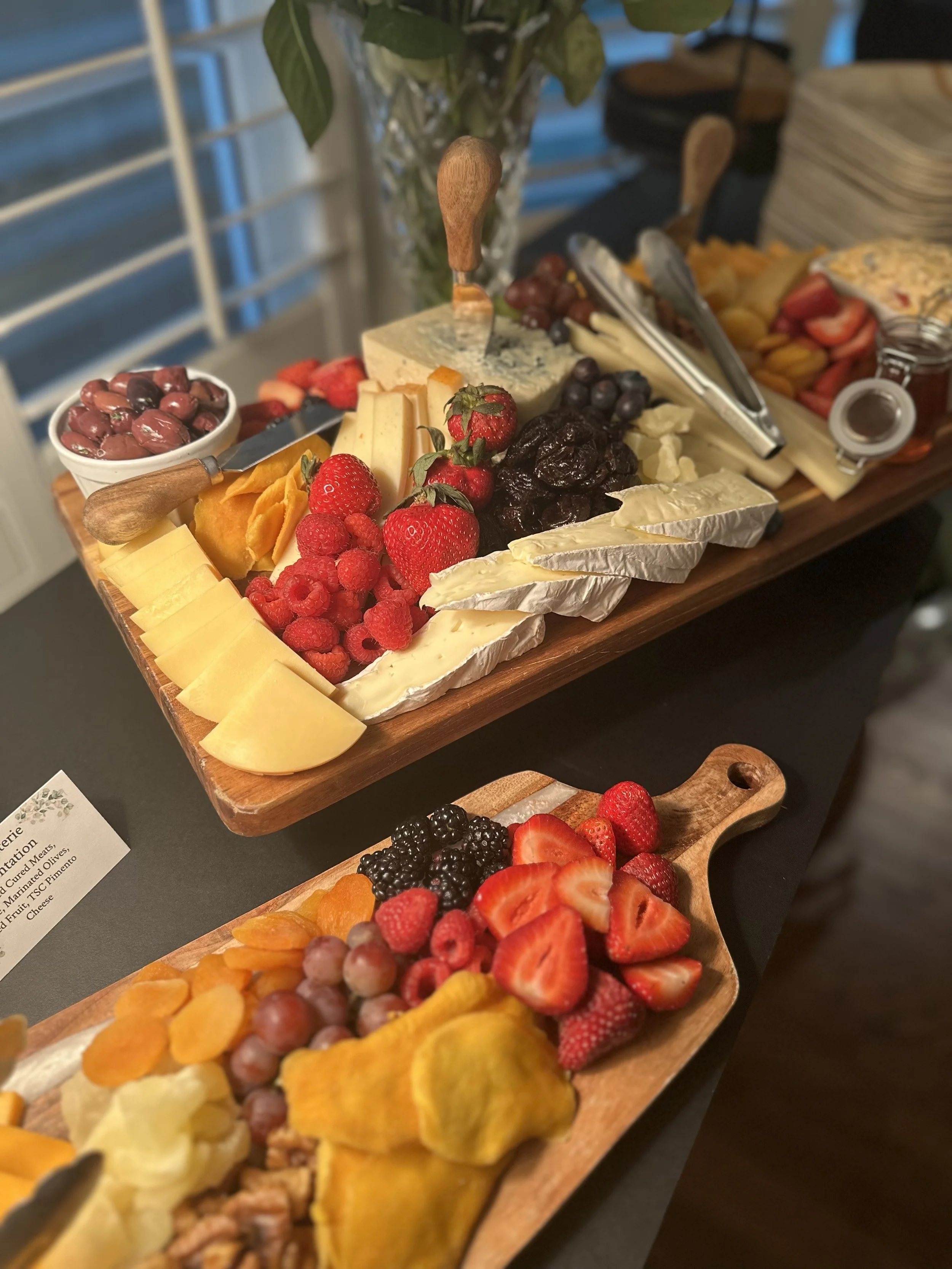 A cheese and fruit platter with various cheeses, fresh strawberries, raspberries, blackberries, grapes, and sliced melon on wooden serving boards, with cheese knives and tongs.