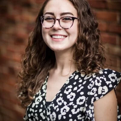A woman with curly hair, glasses, and a black and white printed top, smiling in front of a brick wall.