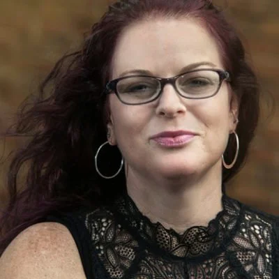 A woman with red hair wearing glasses and hoop earrings, looking at the camera with a slight smile, outdoors with blurred background.