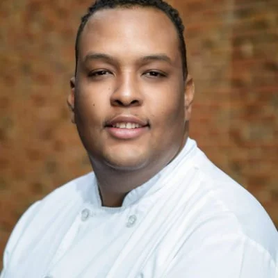 Portrait of a chef in a white uniform with a brick wall background.