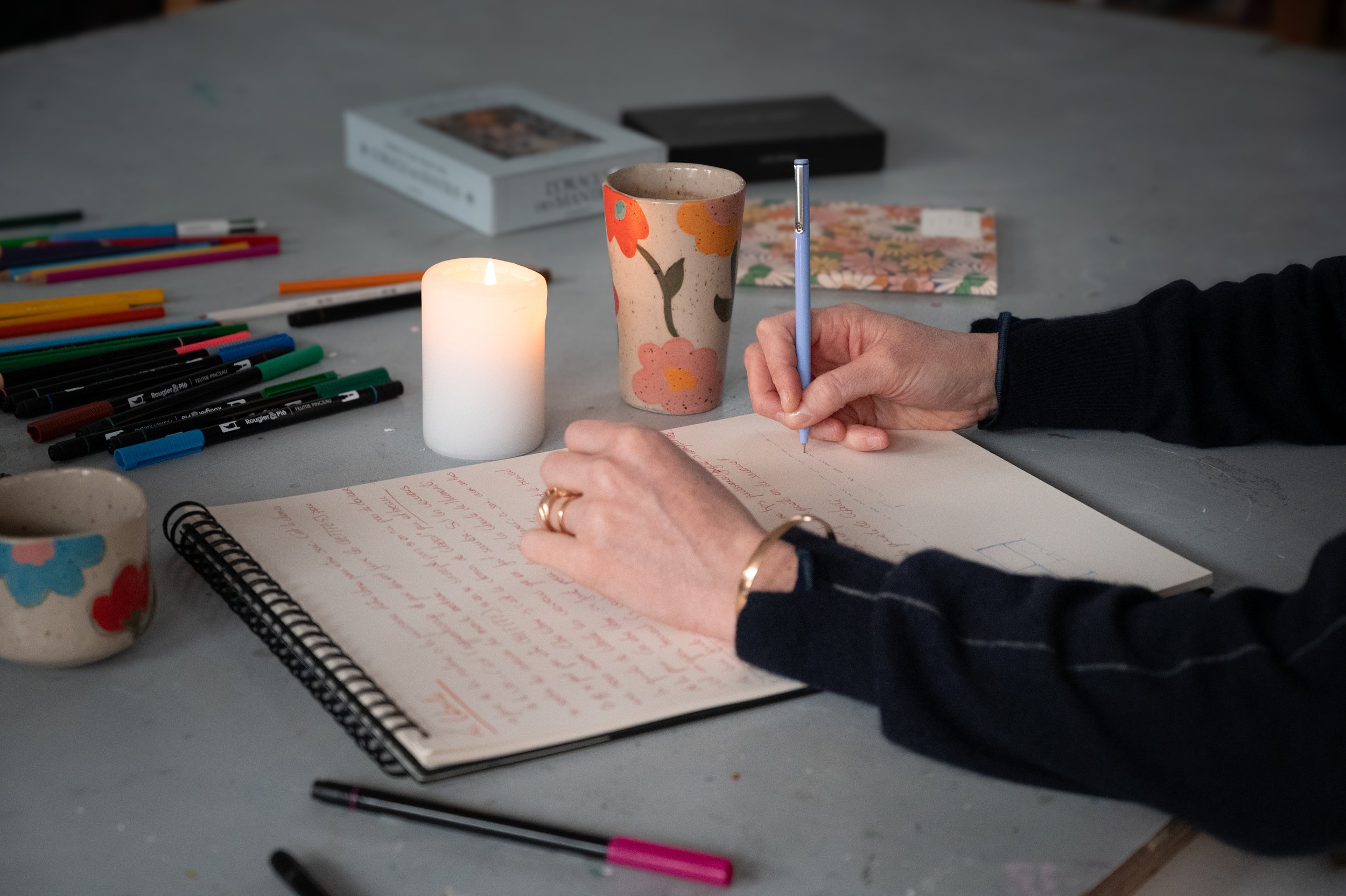 Personne en train d'écrire dans un cahier avec des stylos colorés, assise à une table avec une bougie allumée, une tasse décorative, et plusieurs objets créatifs autour, comme un carnet ou des cartons.