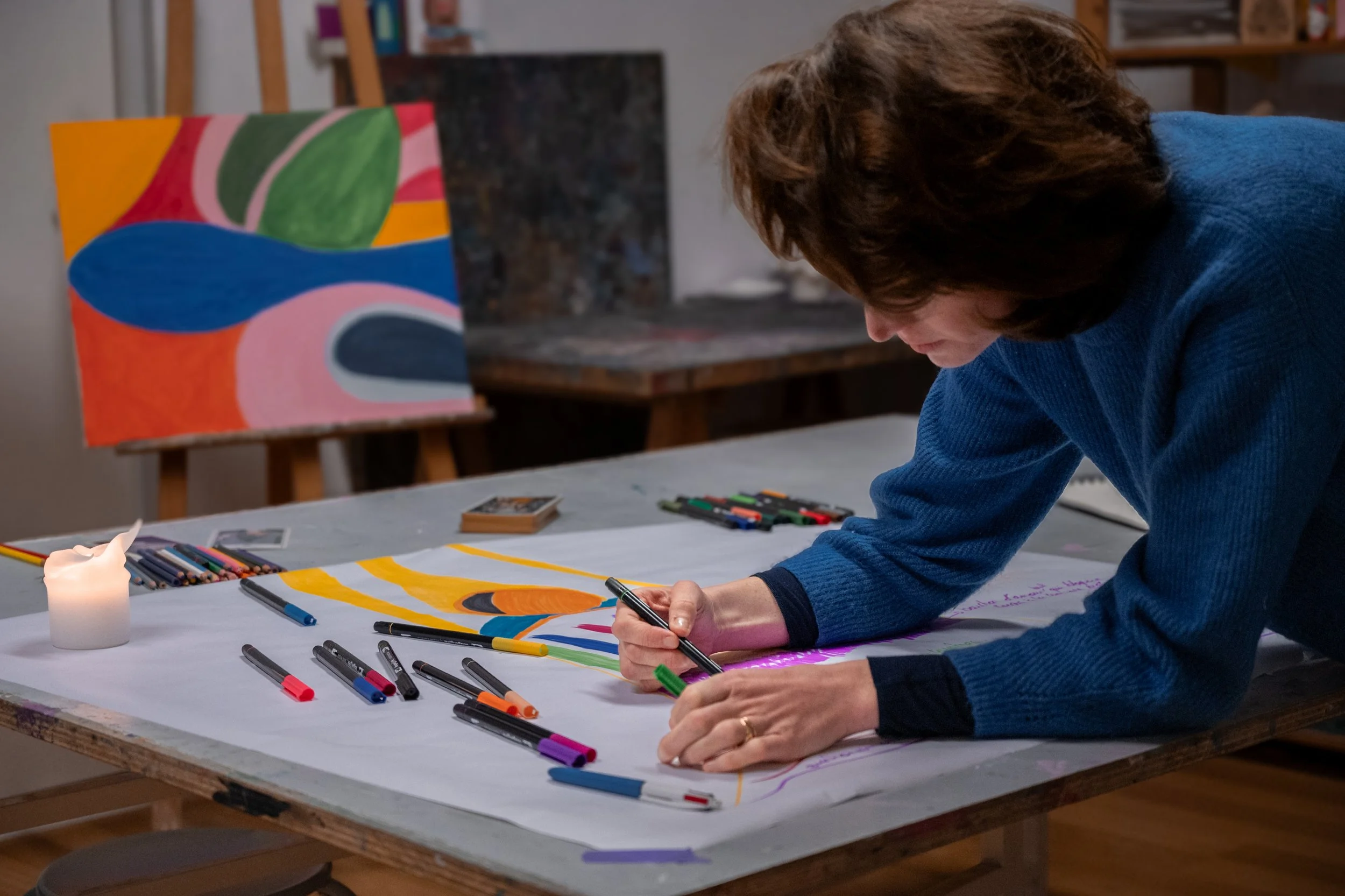 Une femme qui dessine et colore une œuvre d'art abstraite sur une grande feuille de papier, entourée de marqueurs de différentes couleurs, dans un atelier ou studio d'art avec une peinture abstraite en arrière-plan.