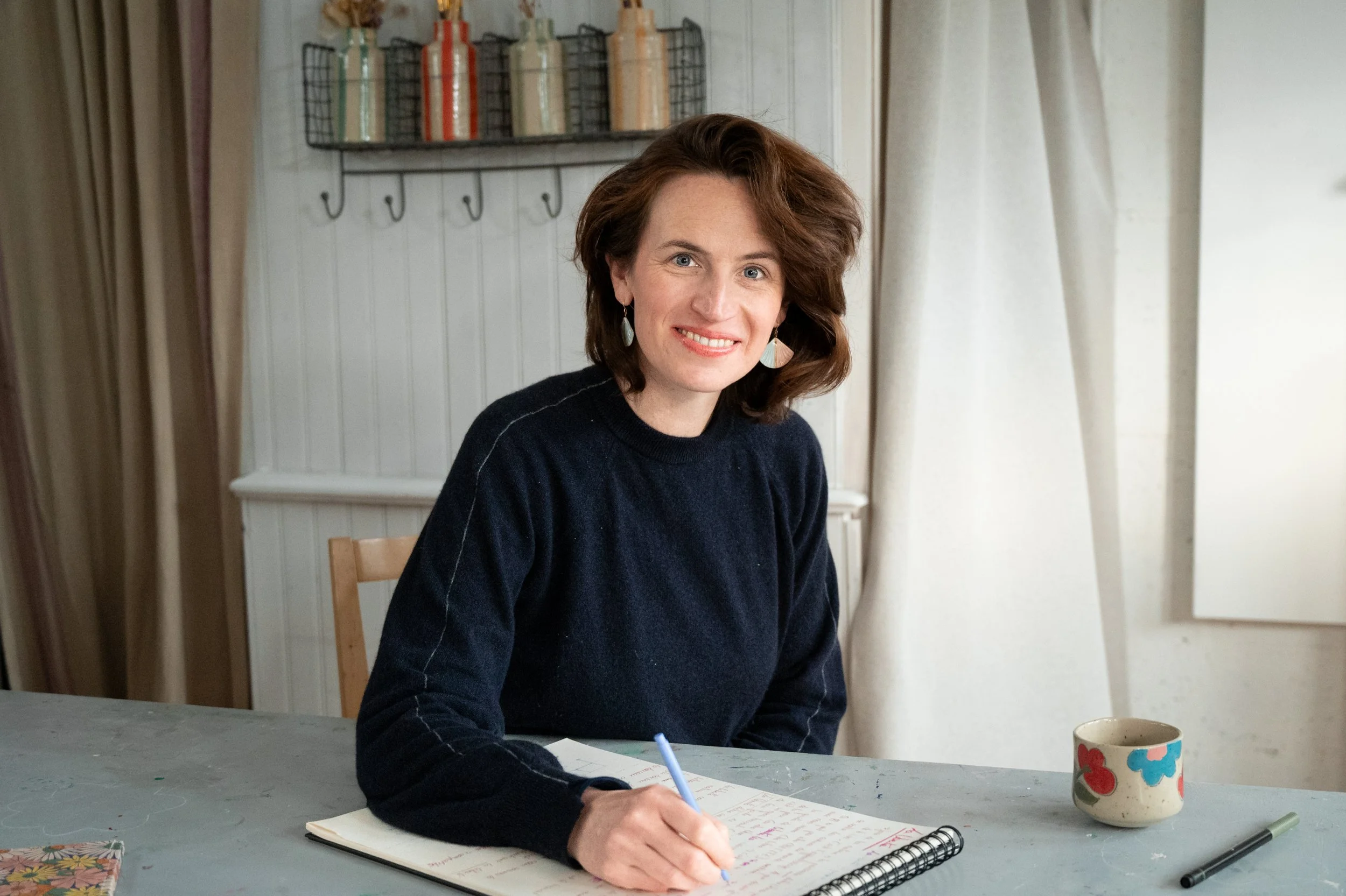 Femme souriante avec cheveux bruns, portant un pull noir, assise à une table avec un carnet, écrivant avec un stylo bleu, dans une cuisine lumineuse, à côté d'une tasse décorative colorée.