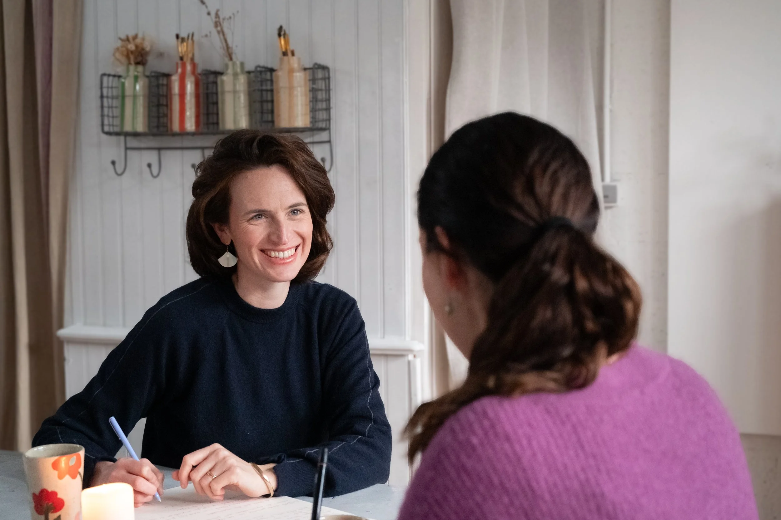 Deux femmes discutent à une table, l'une souriante, avec un décor de cuisine en arrière-plan.