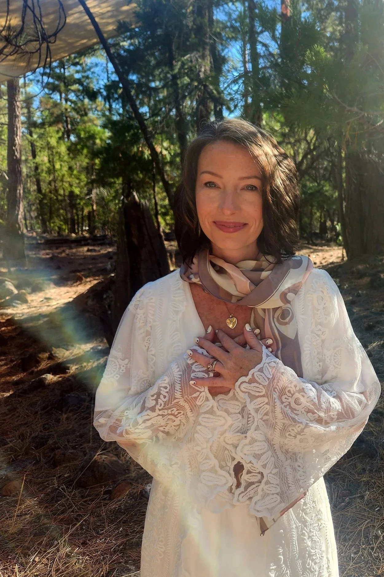 A woman with short brown hair, smiling and wearing a white lace dress, stands in a forest with sunlight filtering through trees, holding her hands over her chest, with a scarf around her neck and a gold heart necklace.