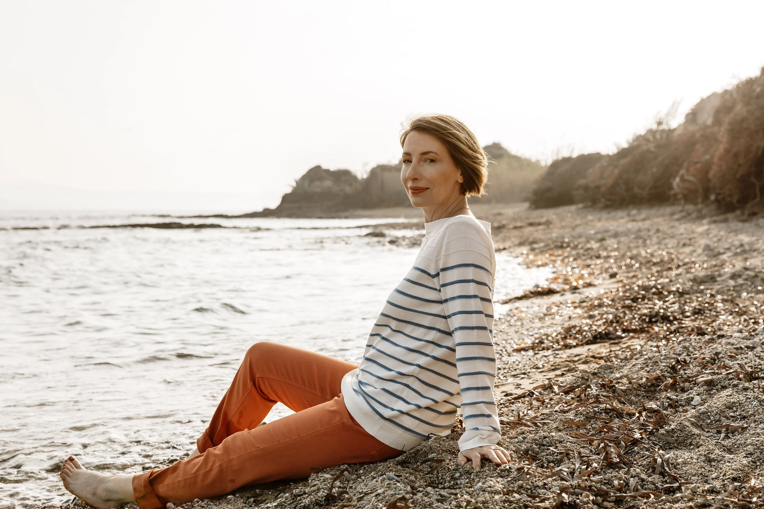 RaChel Benyair, wearing a white and navy striped sweater and orange pants, sitting barefoot on a pebbled beach near the water, with a rocky headland and trees in the background during sunset.