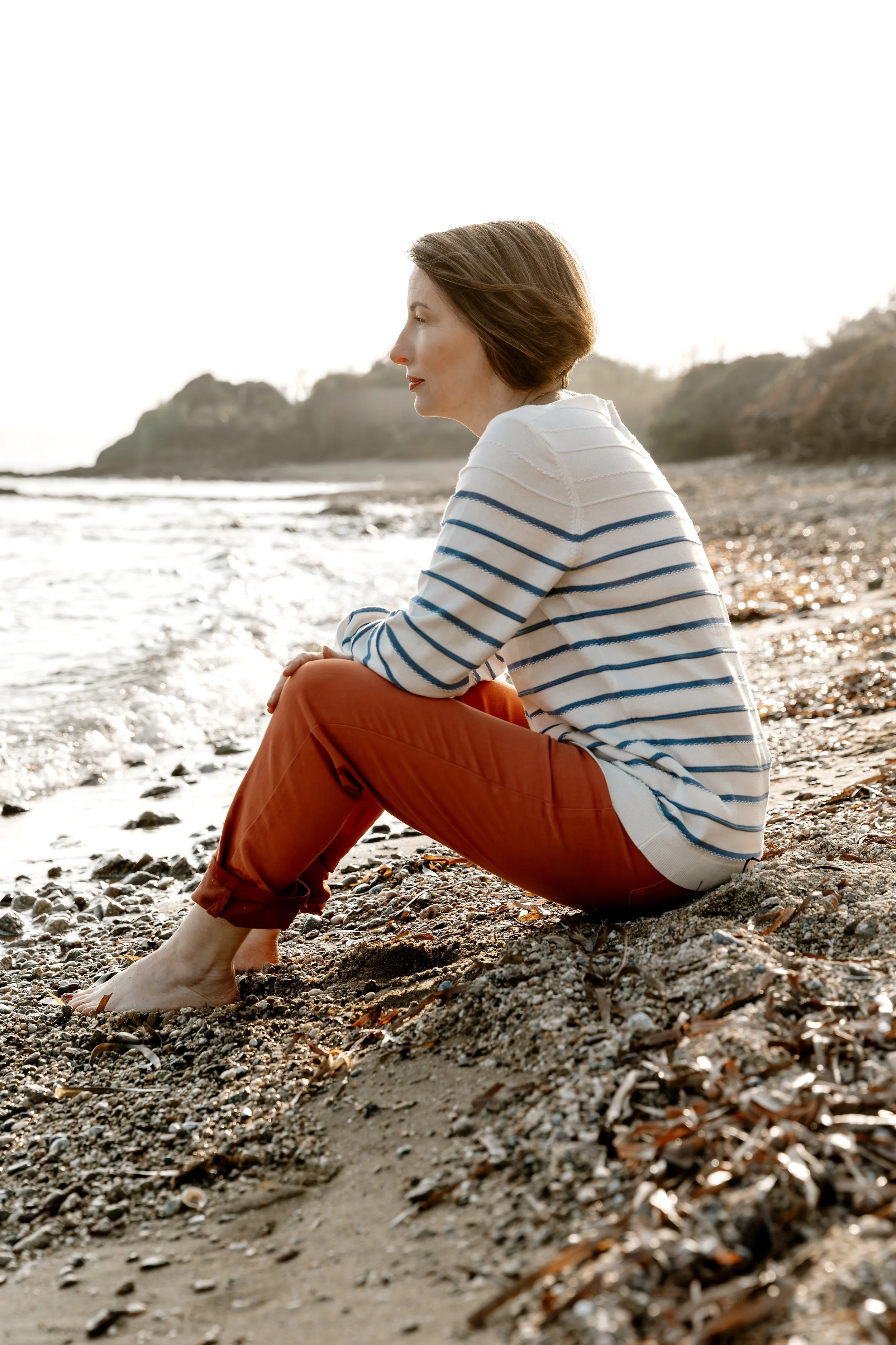A woman with short brown hair, wearing a striped sweater and red pants, sits on a beach with her feet in the sand, looking peacefully toward the water during sunset or sunrise.