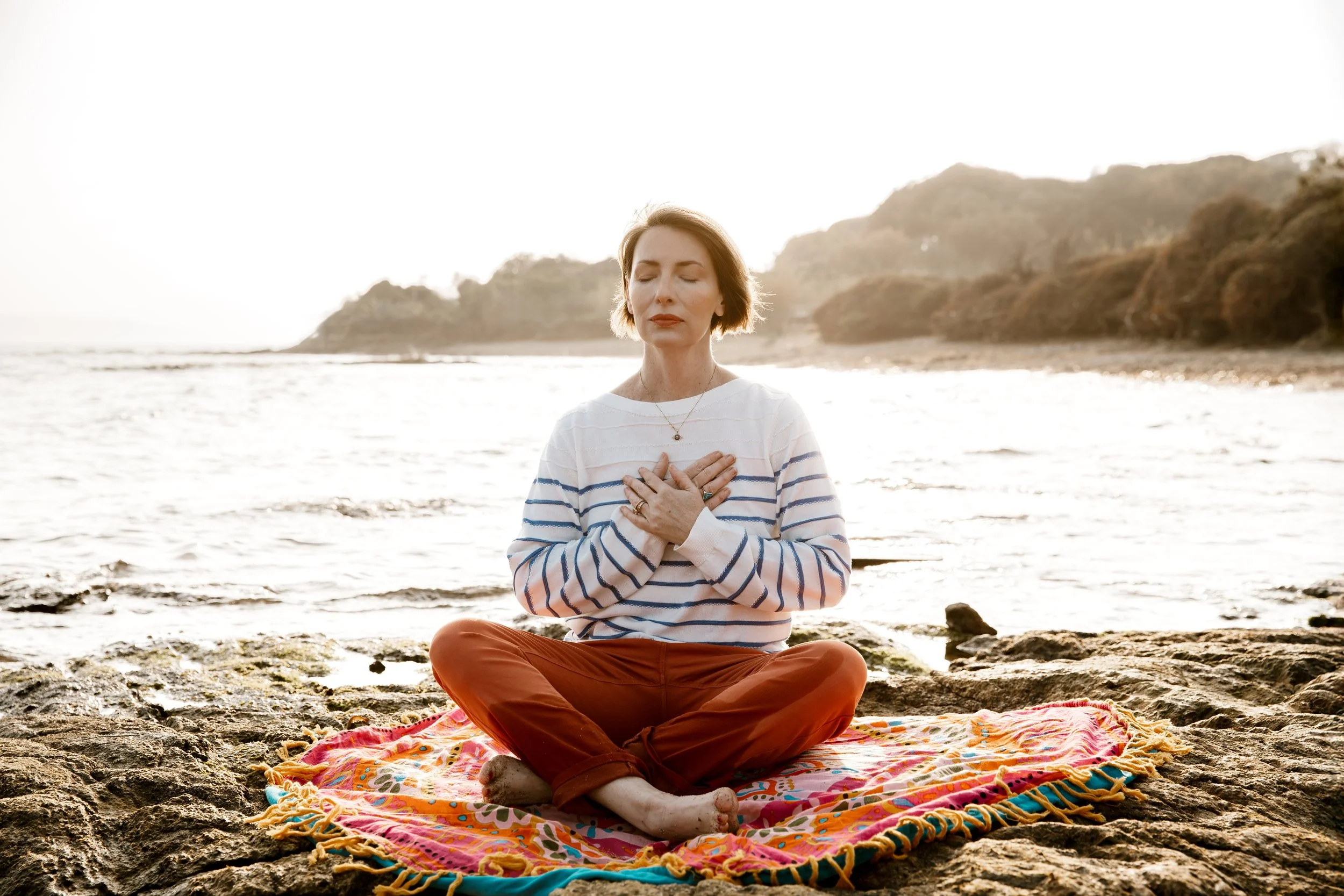 Woman meditating on a colorful blanket by the beach with her hands on her chest, eyes closed, during sunset.