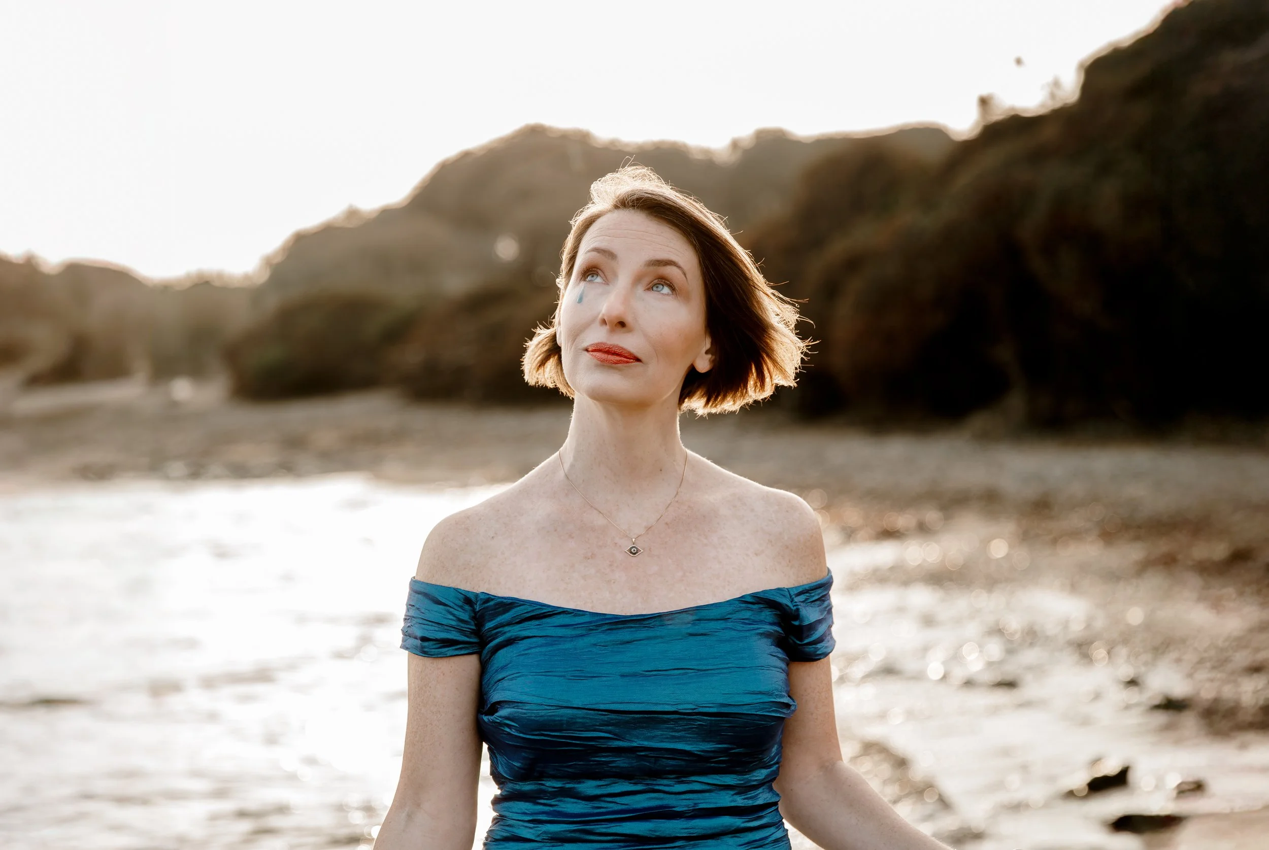 A woman in a blue off-shoulder dress standing on a beach during sunset, looking up with a contemplative expression.