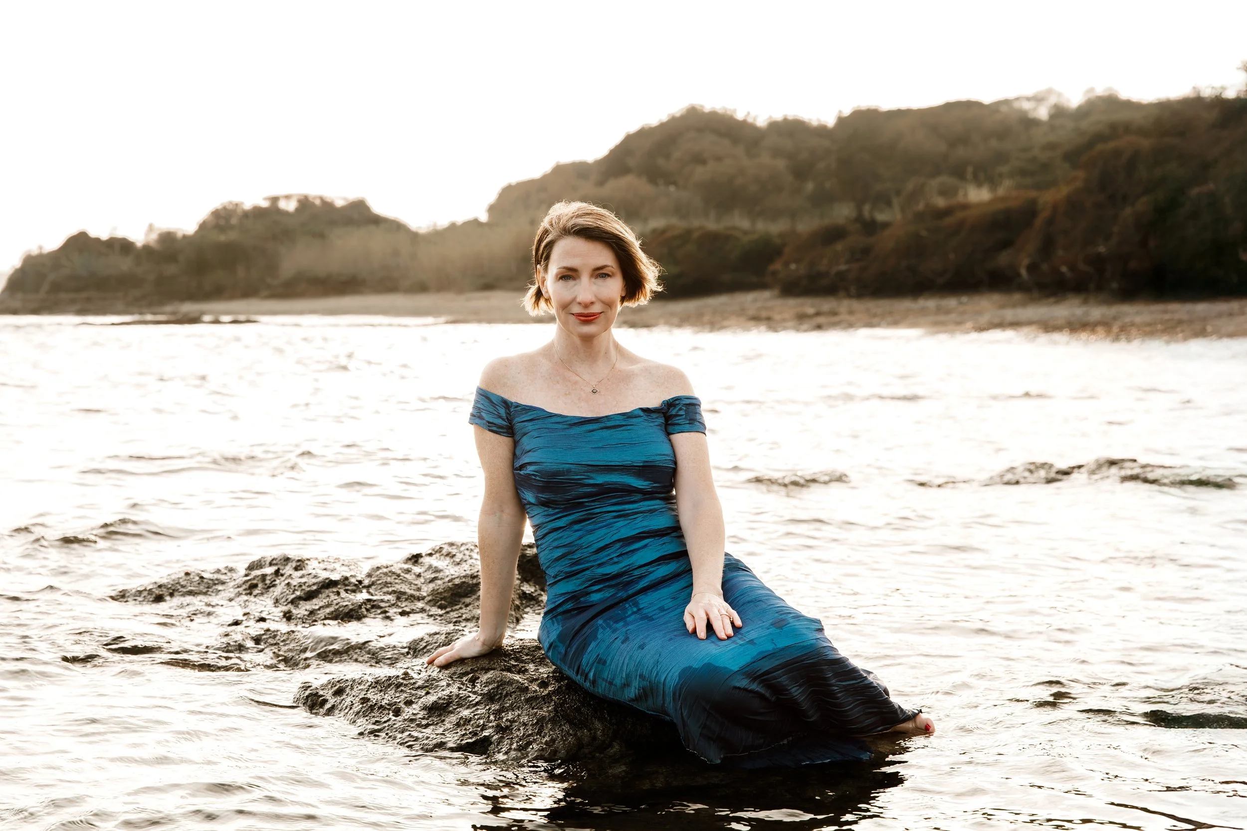 A woman in a blue off-shoulder dress sitting on a rock in the water at a beach during sunset.