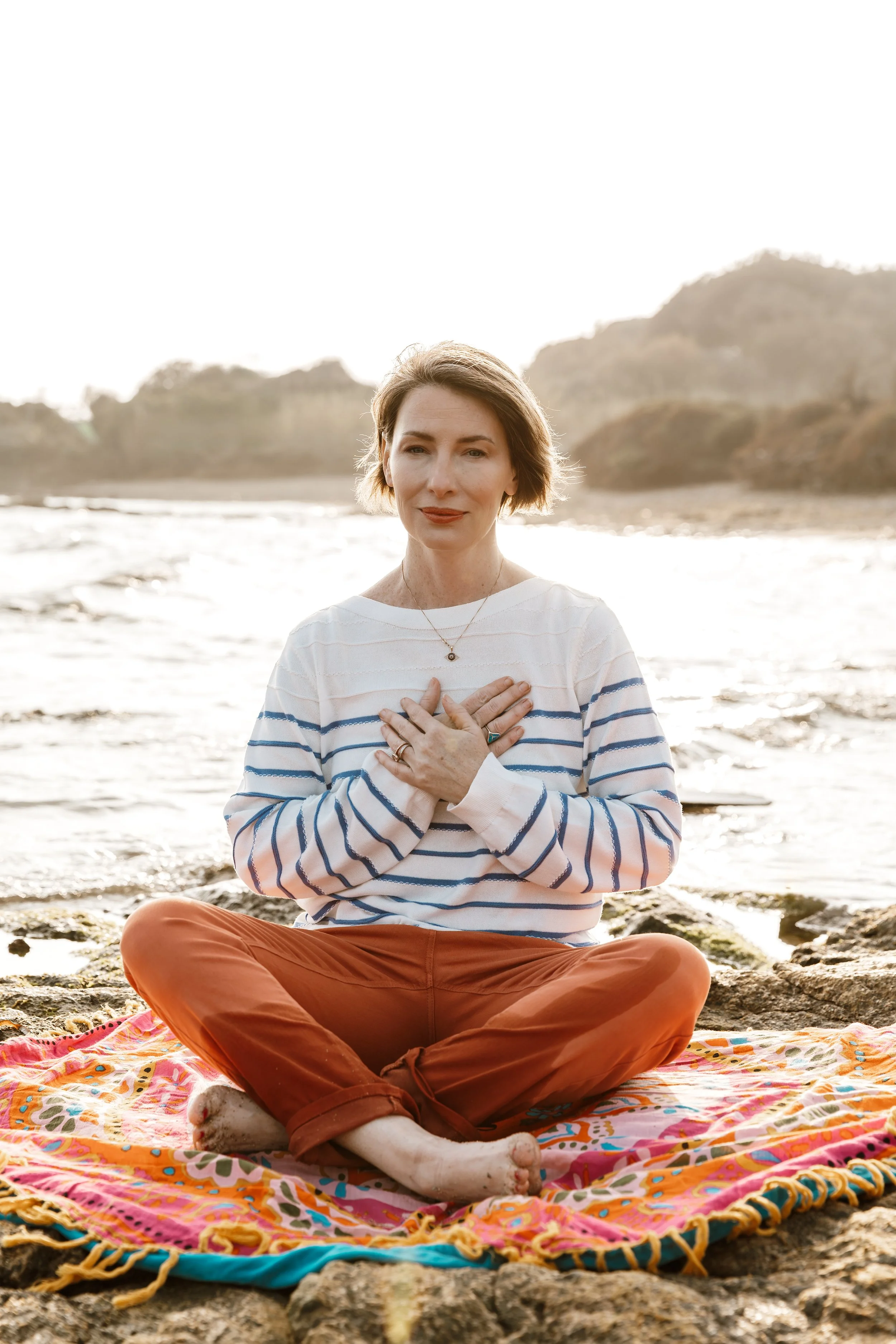 RaChel sitting cross-legged on a colorful blanket at the beach, with her hands on her chest, eyes closed, and a peaceful expression.