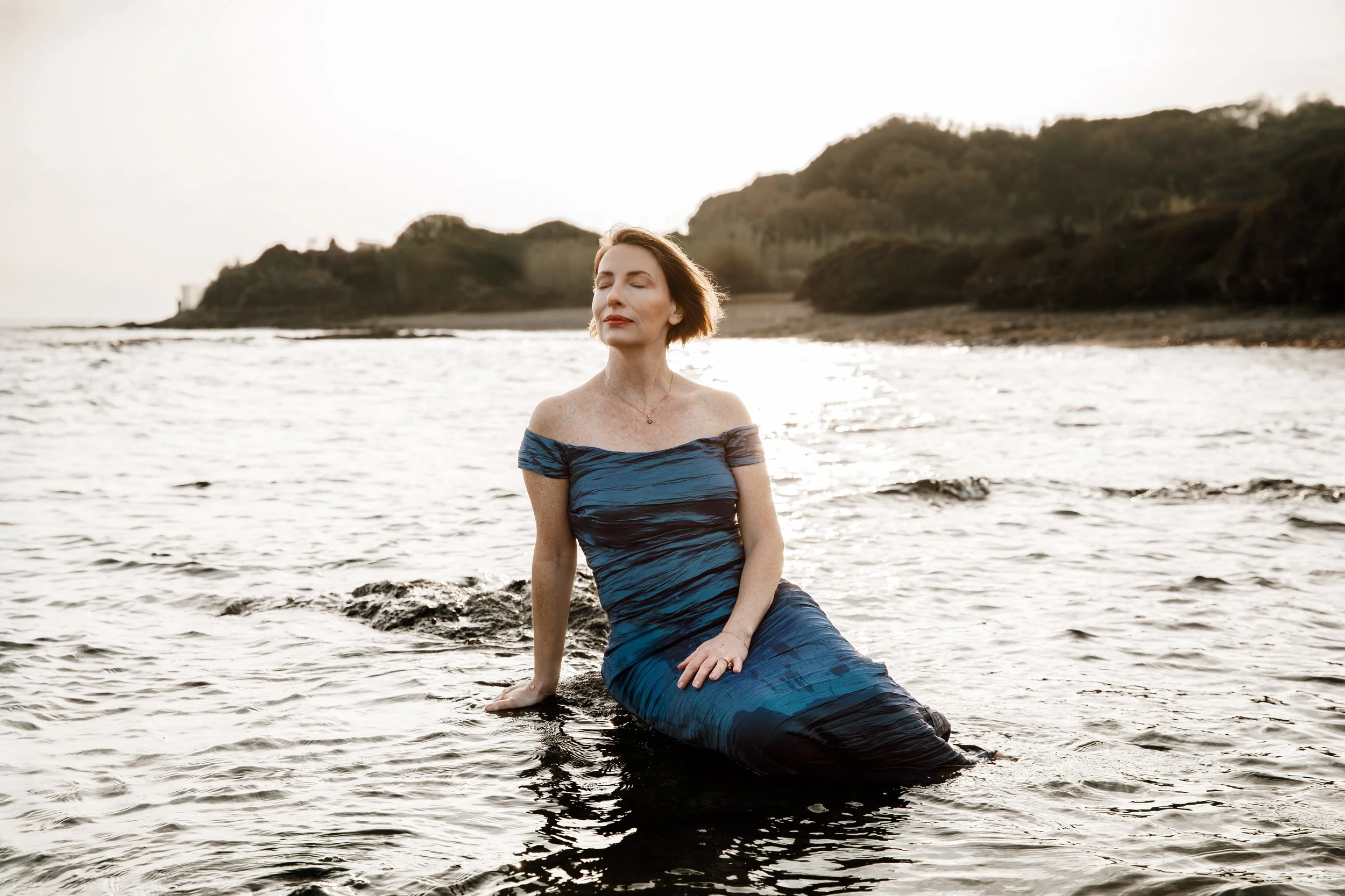 A woman in a strapless, blue and black dress sitting in shallow water at a beach, with her eyes closed and a peaceful expression, during sunset or sunrise.