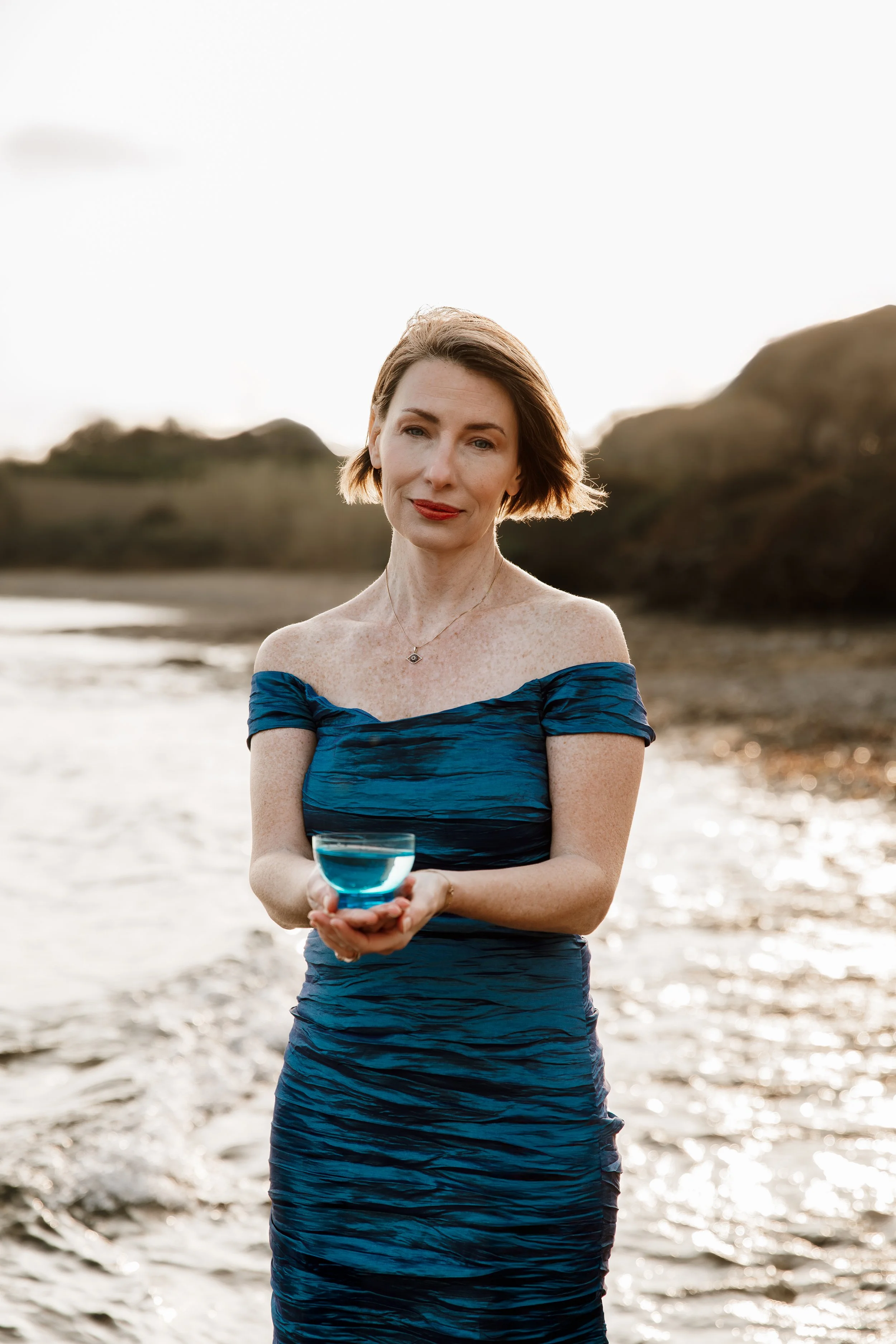 A woman in a blue off-shoulder dress holding a glass of water on a beach at sunset.
