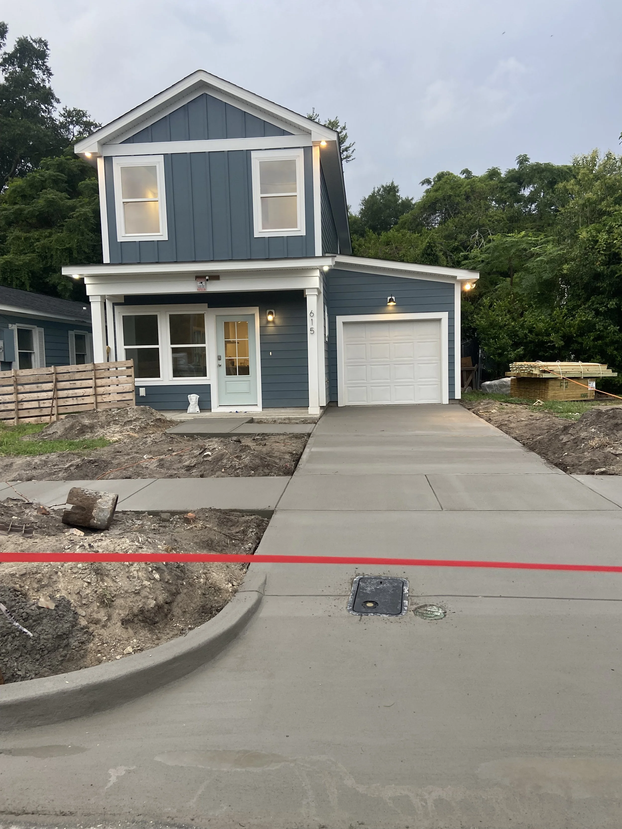 Newly constructed two-story blue house with white trim, a garage, and a concrete driveway, surrounded by construction site dirt and a wooden fence, with greenery and trees in the background.