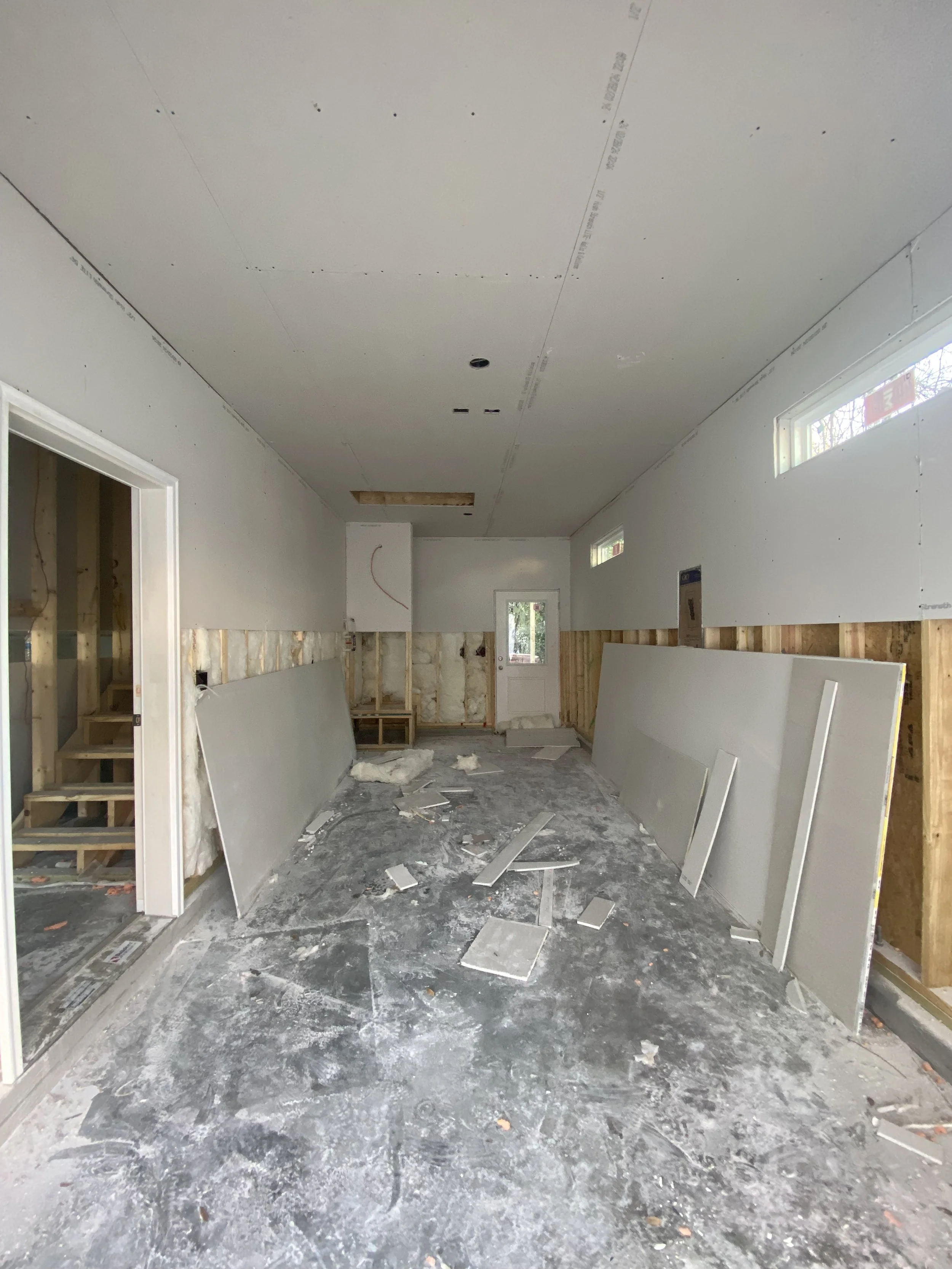 Interior view of a room under construction with drywall panels leaning against the walls, construction debris on the floor, and a doorway leading to another room.