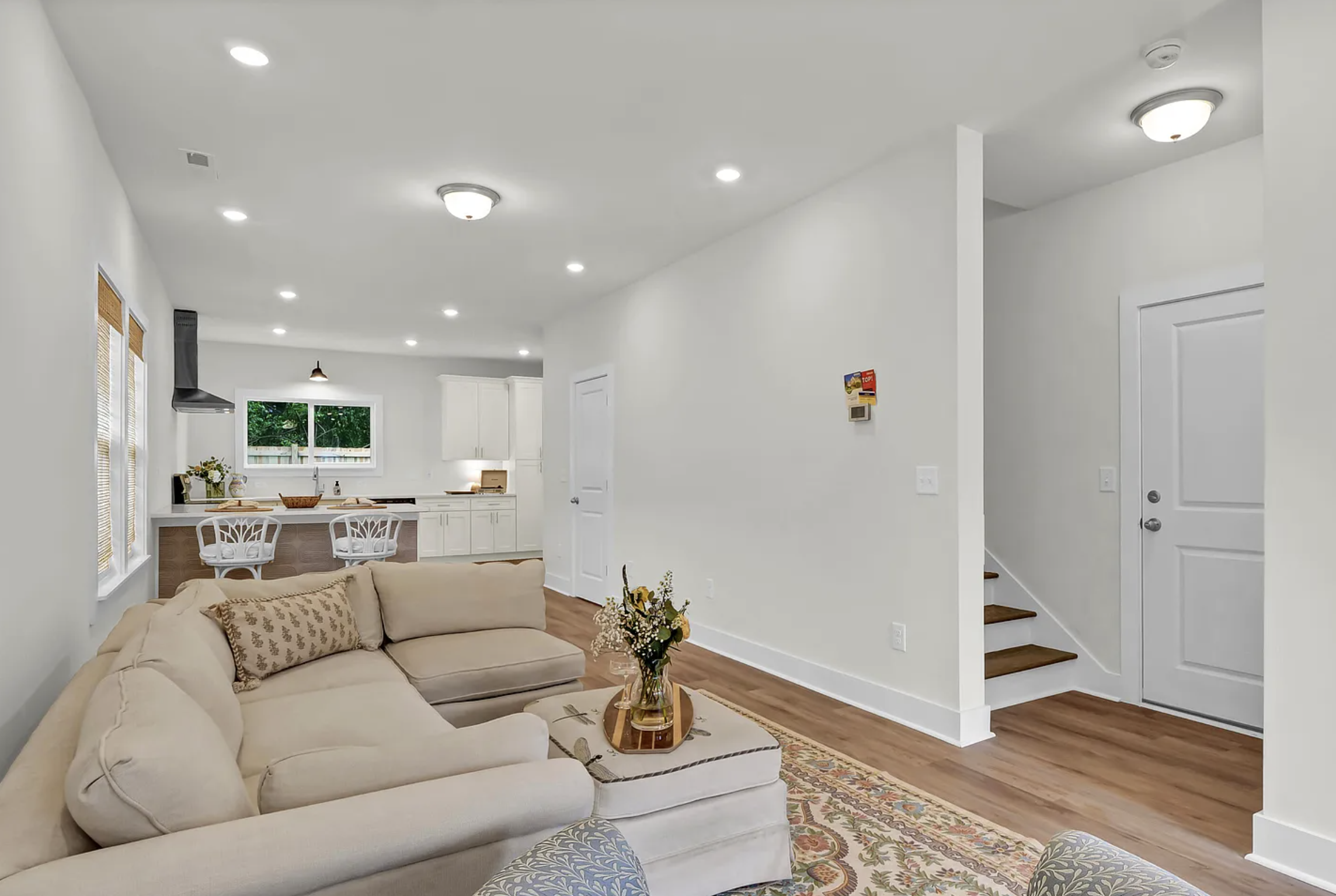 A living room with a beige sectional sofa, a coffee table with a flower arrangement, hardwood floors, white walls, and a kitchen in the background with white cabinets, a window, and a breakfast bar.