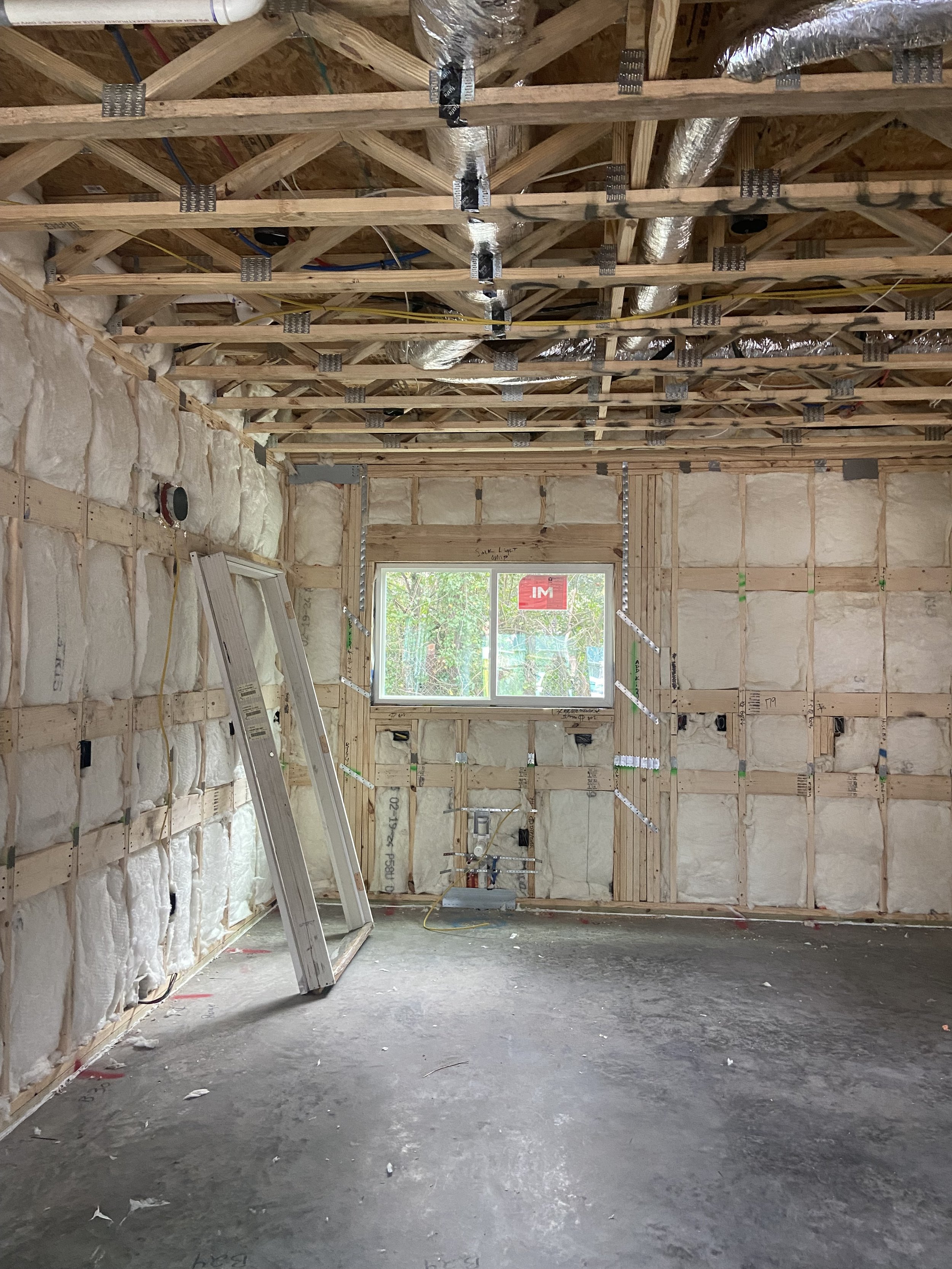 Interior view of a house under construction, showing wooden framing, insulation, electrical wiring, and a window.