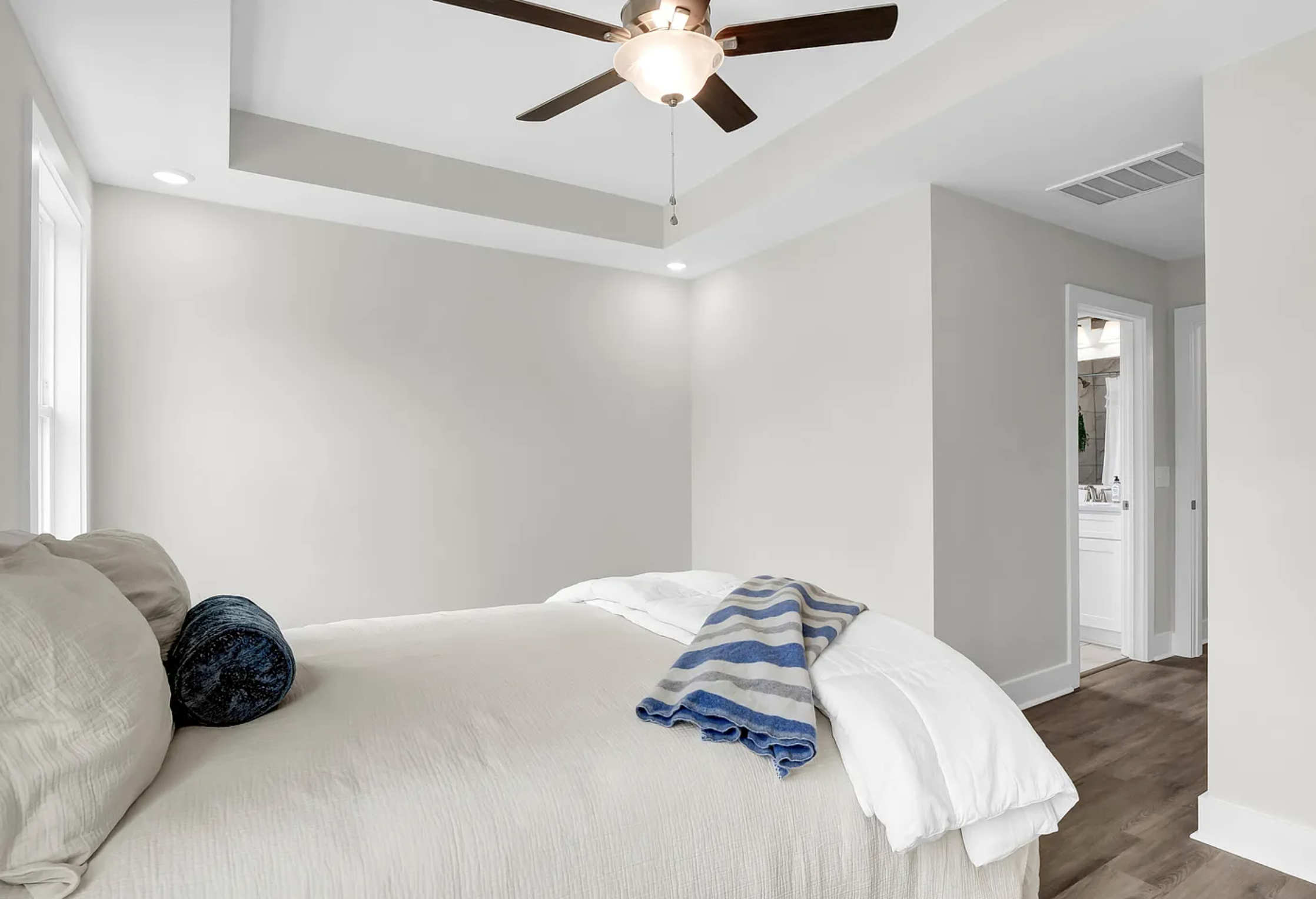 A bedroom with a neatly made bed with beige and white bedding, a striped blue and white blanket, and a navy pillow. The room is painted in light neutral colors, has wood flooring, and a ceiling fan with dark blades and a central light. There is a doorway leading to a bathroom visible in the background.