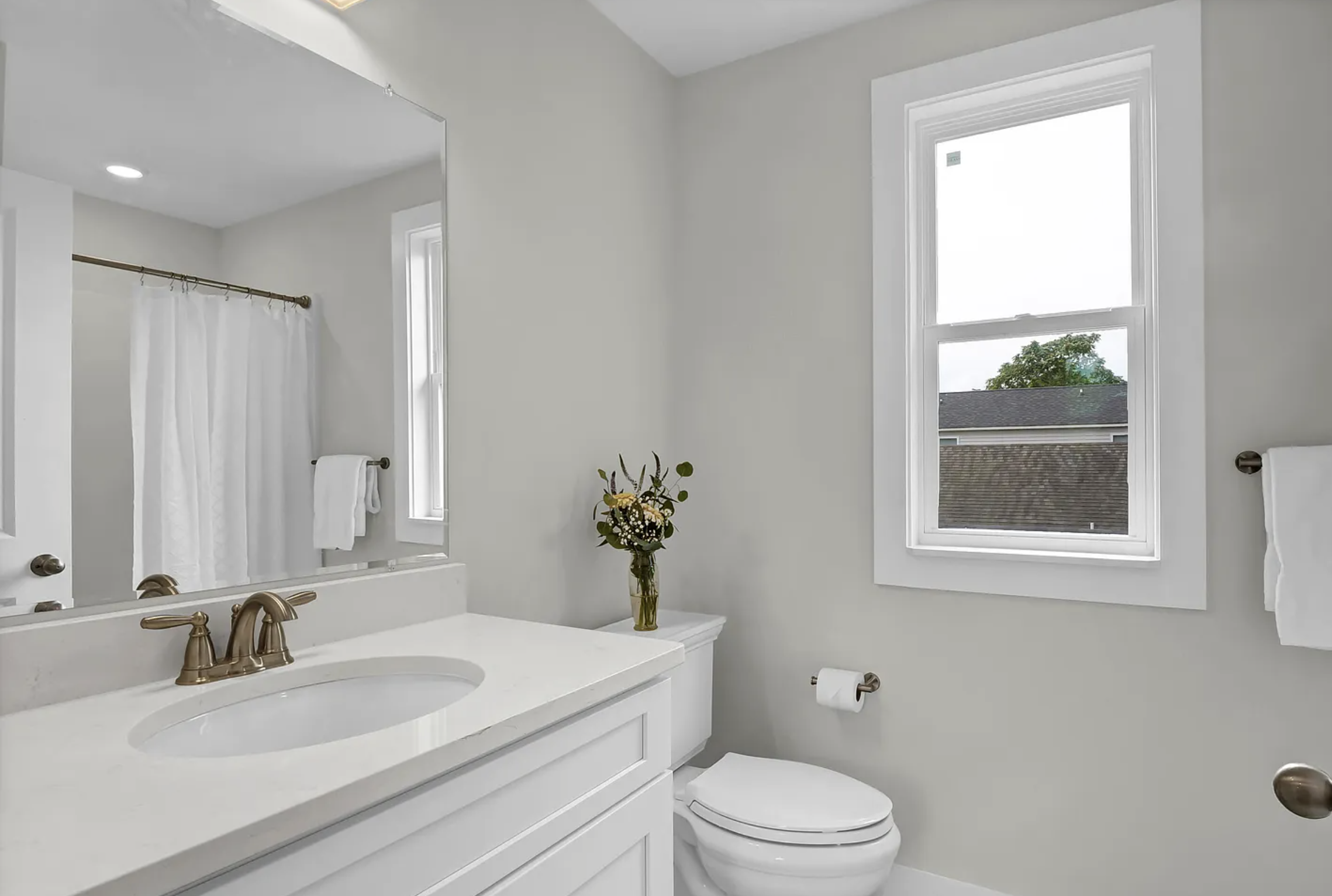 Bathroom with white vanity, sink, mirror, window, and a vase with flowers.