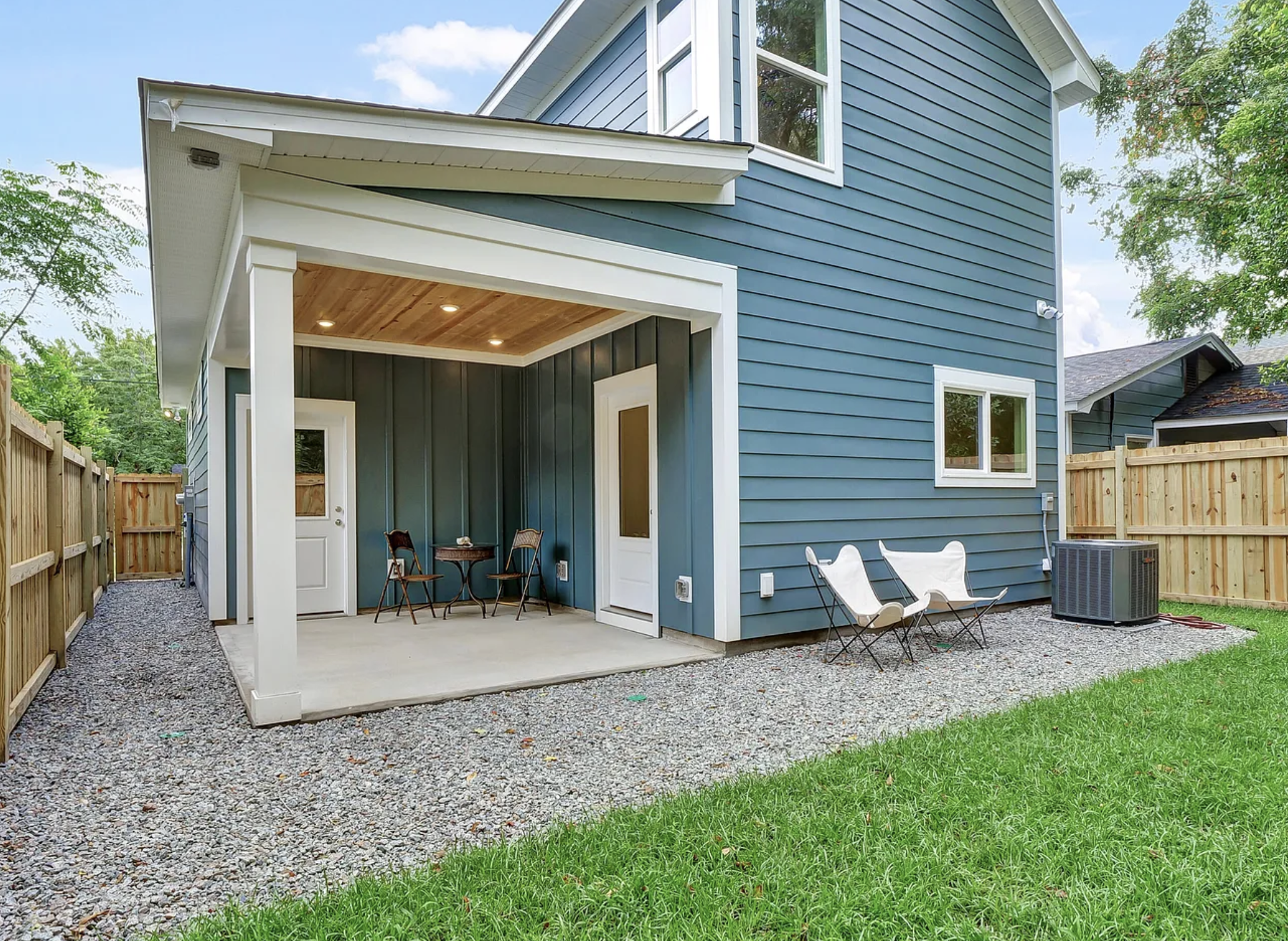 Backyard with blue house, covered patio with seating, gravel ground, green lawn, and wooden fence.