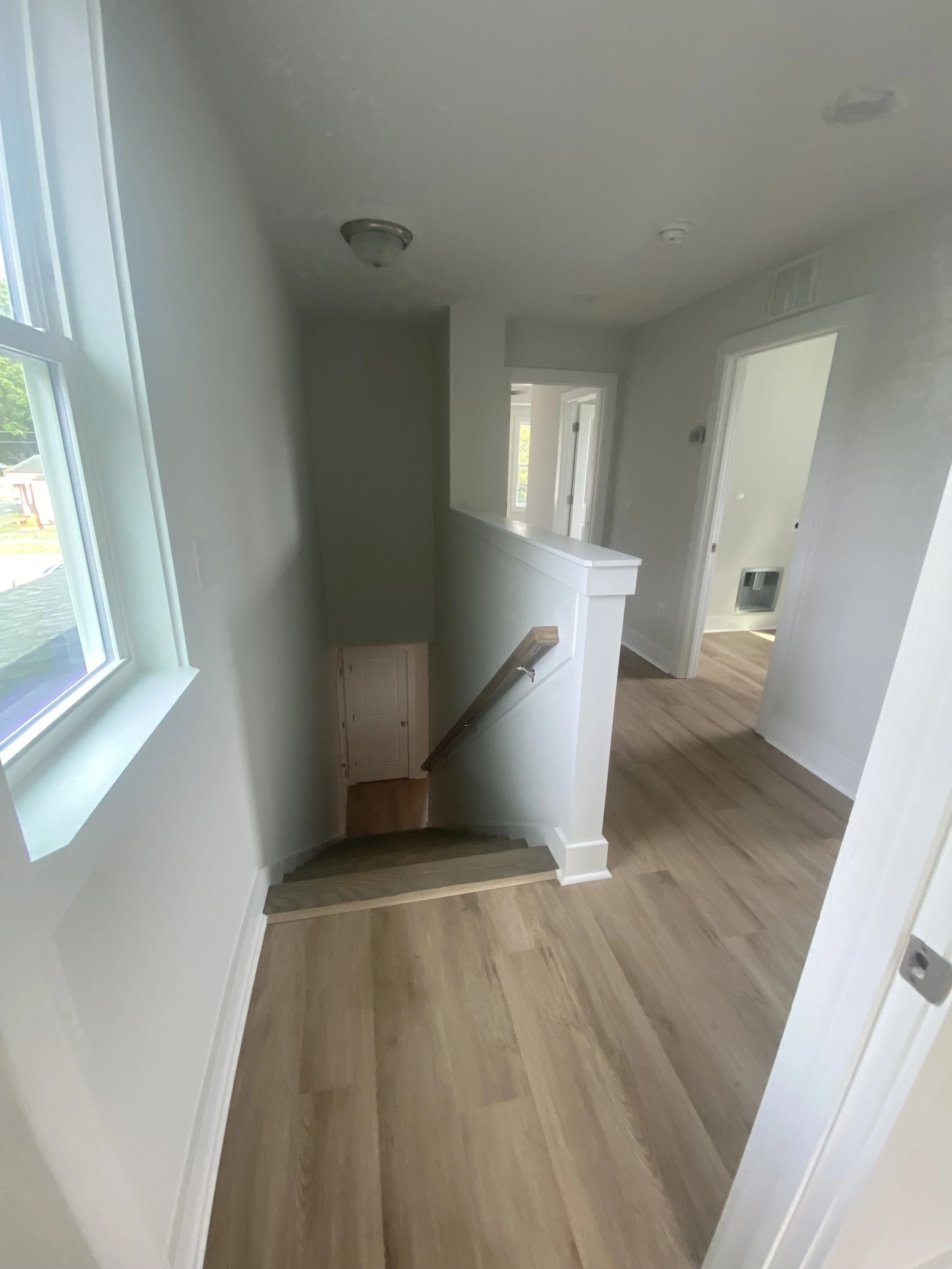 View of a hallway in a house with wooden flooring, a staircase leading down, white walls, a window on the left, and doors leading to other rooms.