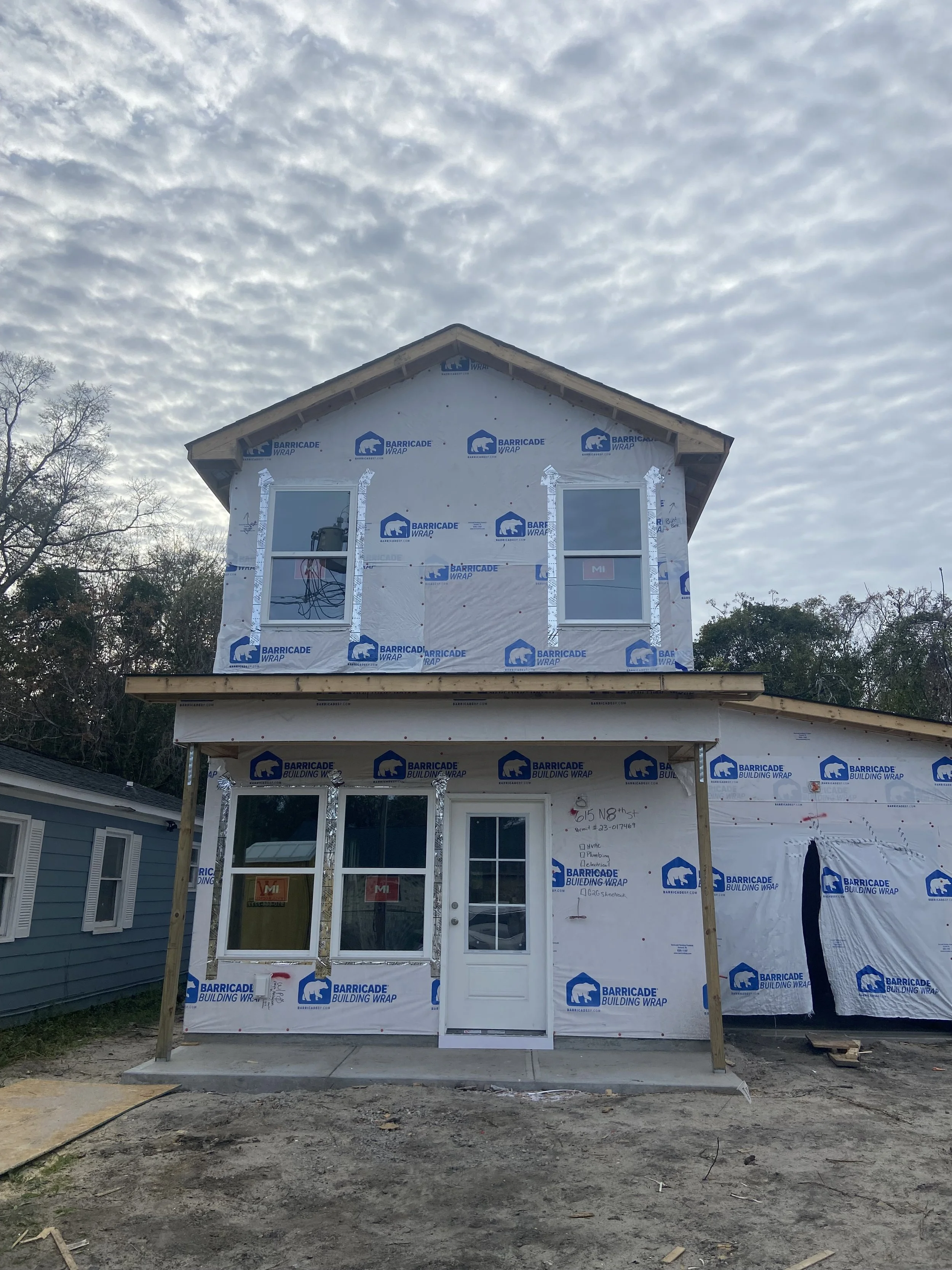 Two-story house under construction with building wrap and unfinished windows, located next to a smaller light blue house, with cloudy sky overhead.