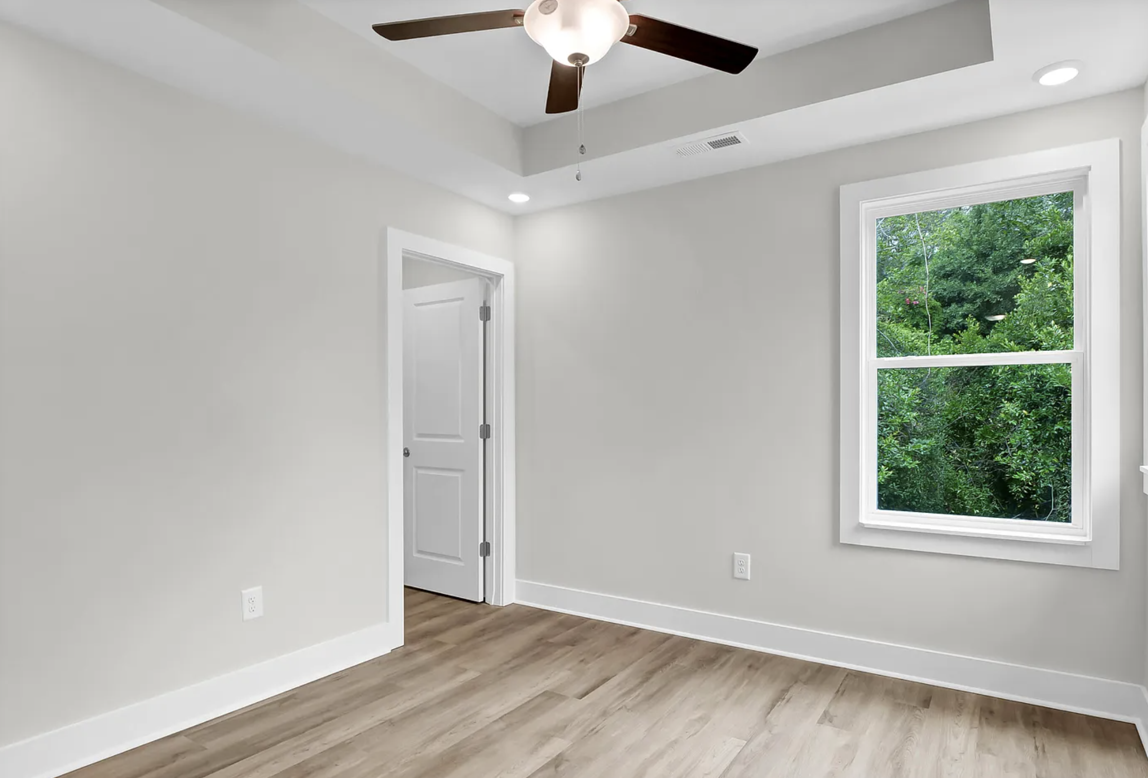 Empty room with light-colored walls, a window showing green trees outside, a ceiling fan, and hardwood flooring.