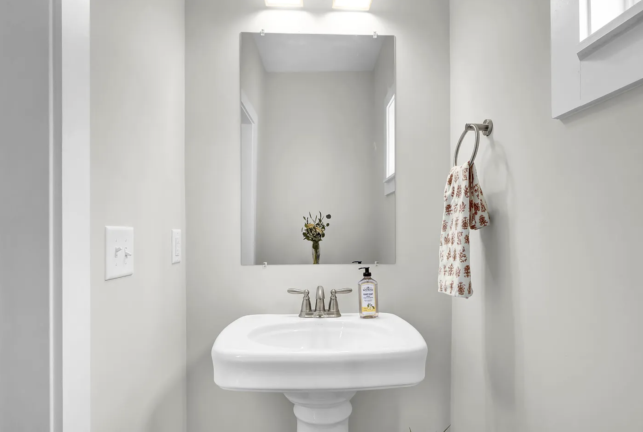 Minimalist bathroom with white walls, a pedestal sink, a large mirror, a soap dispenser, a towel ring with a patterned towel, a small window, and a vase with flowers in the mirror reflection.