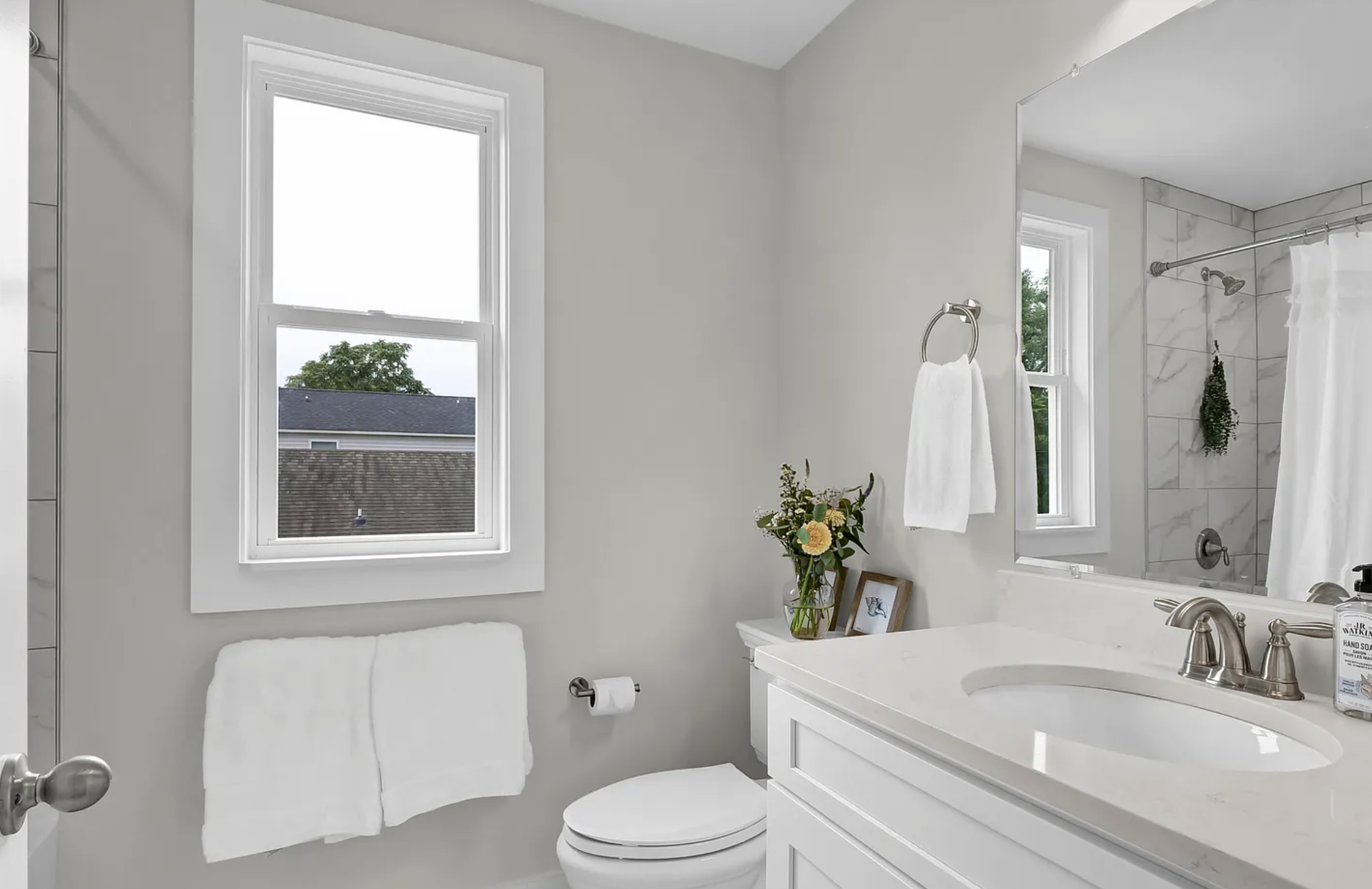 Bright bathroom with a window, white walls, a white toilet, a white vanity with a sink, a large mirror, and a shower with a white curtain and hanging plants.