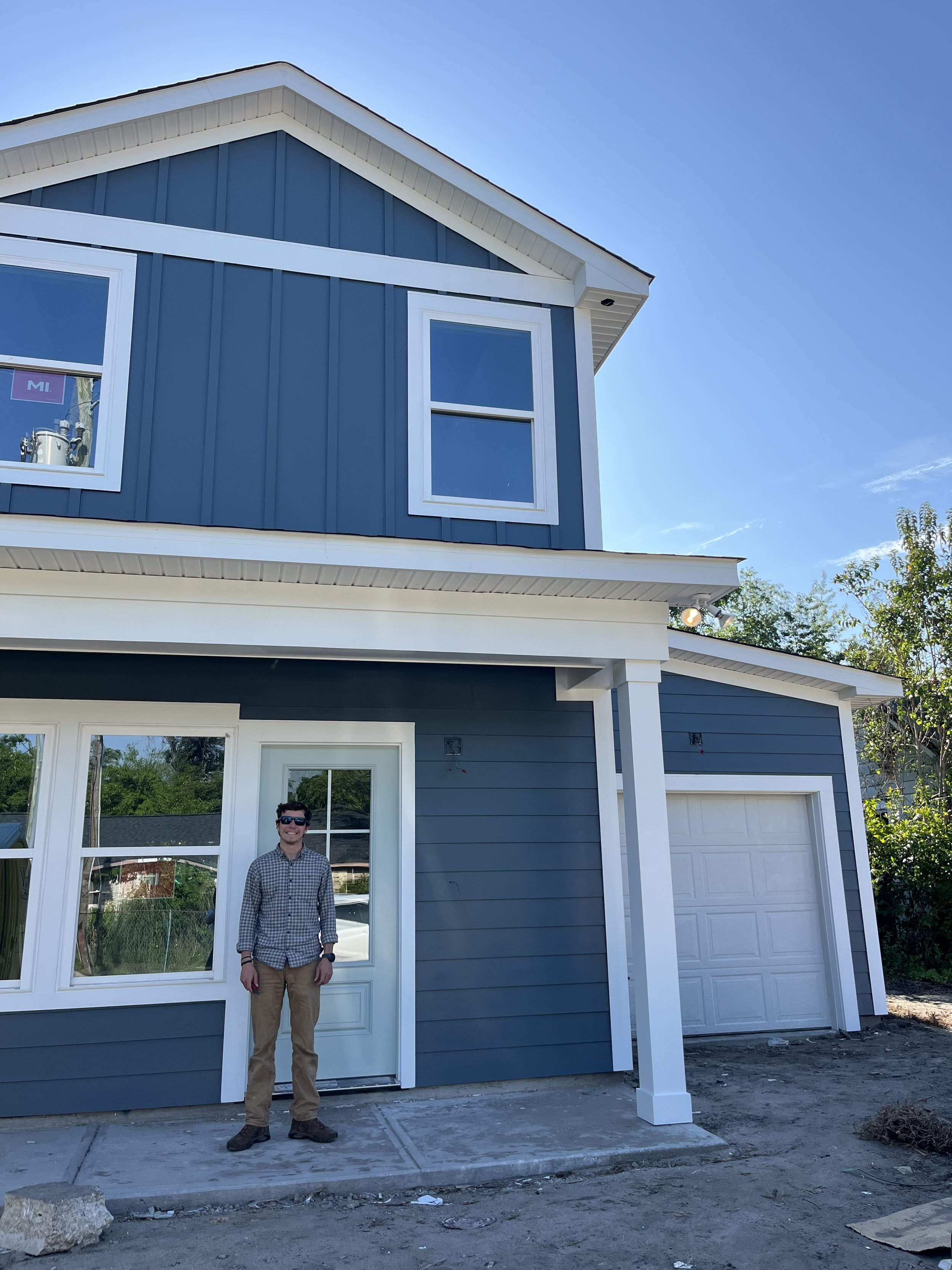 A man standing in front of a newly constructed two-story house with blue siding, white trim, and a closed garage door, under a clear blue sky.